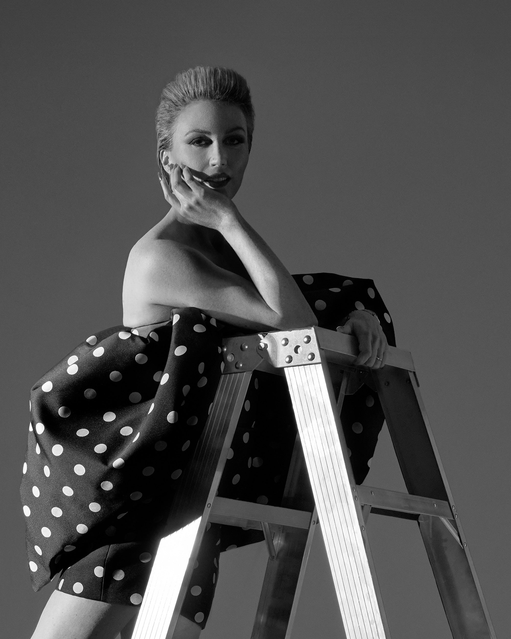 Black and white portrait of a woman leaning on a metal ladder wearing a polka dot dress against a plain background