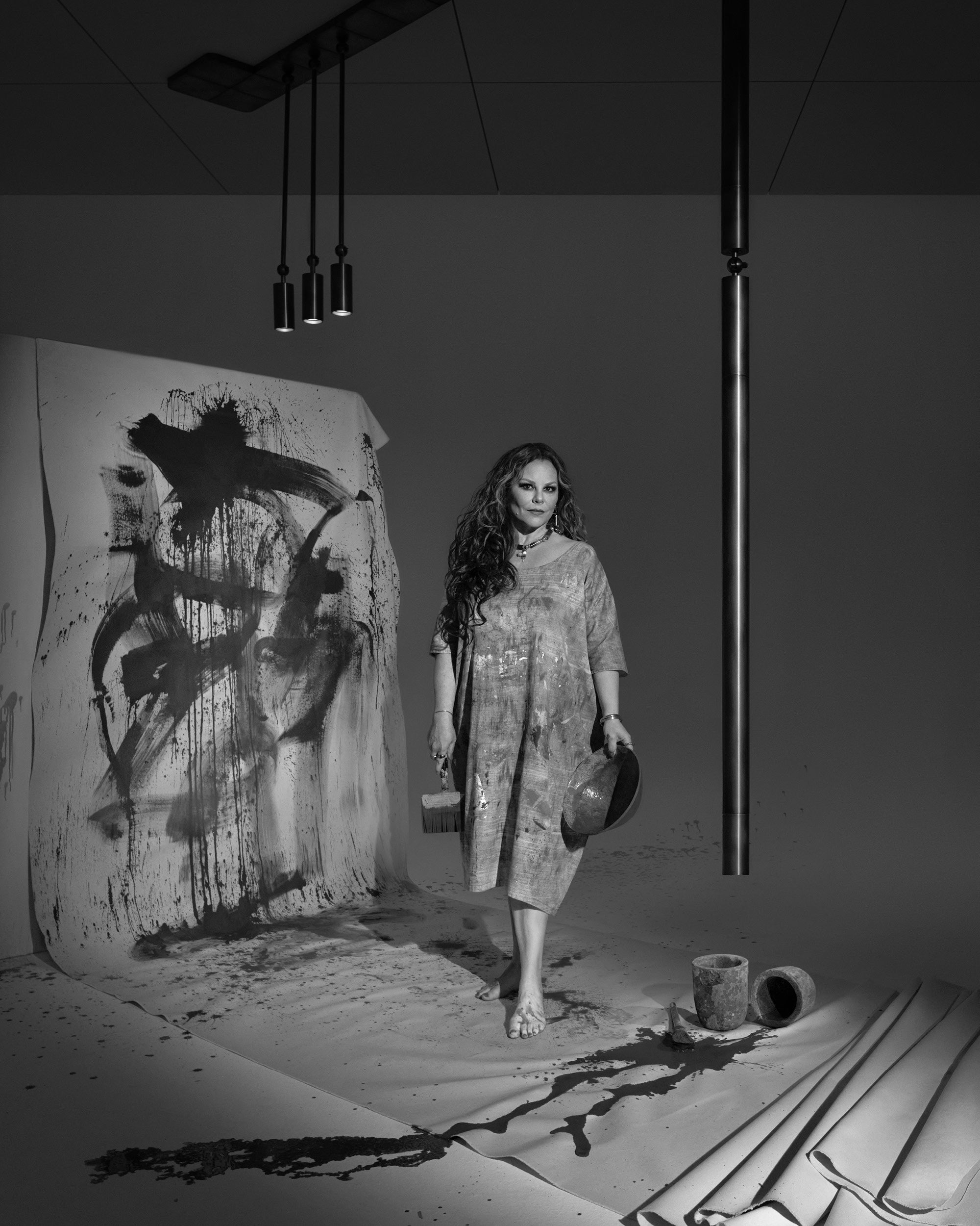 Black and white image of a barefoot woman standing beside abstract ink painting with spilled paint on the floor beneath hanging lights
