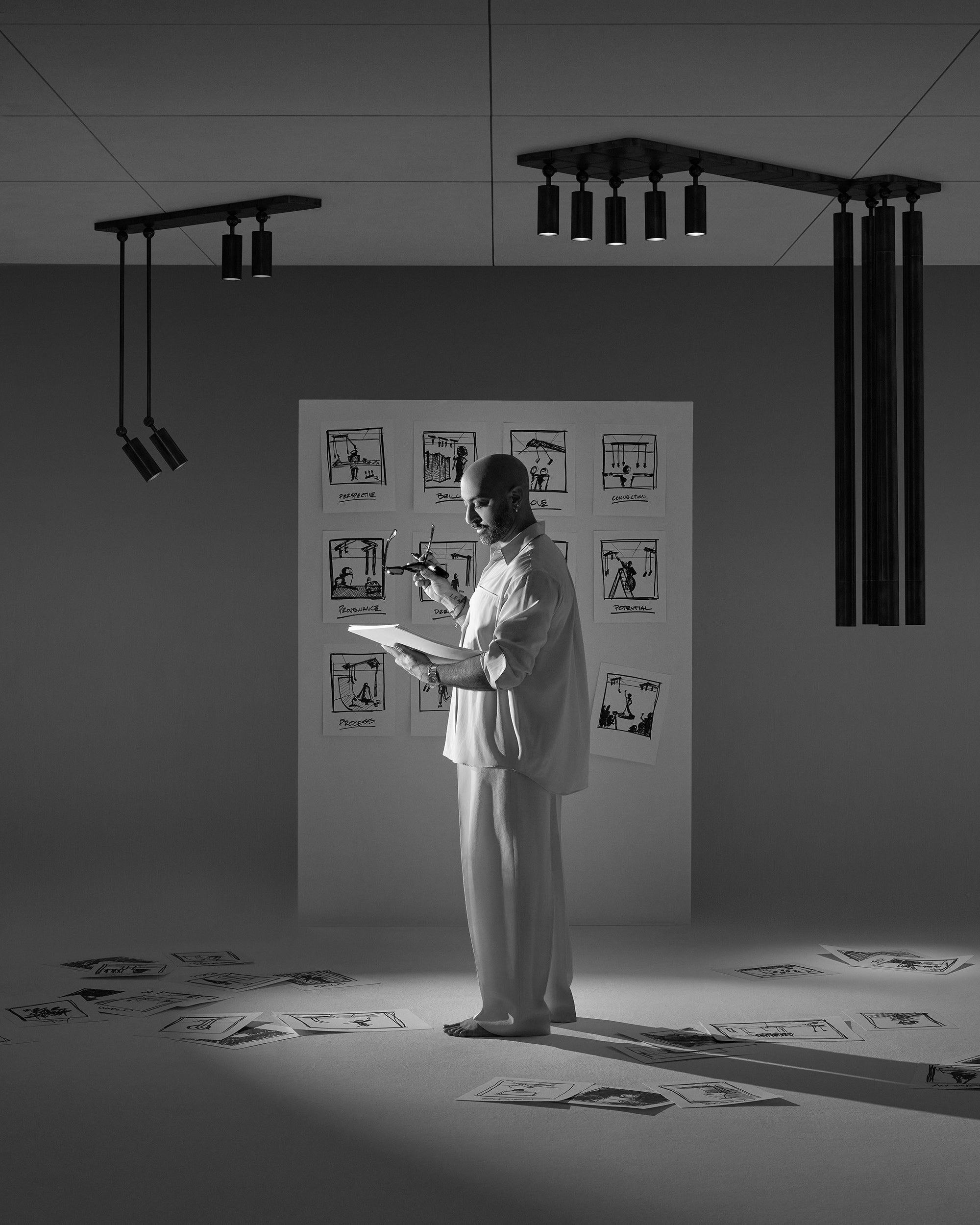 Black and white image of a man in light clothing holding a sketchbook and angled drawings pinned and scattered on floor and wall
