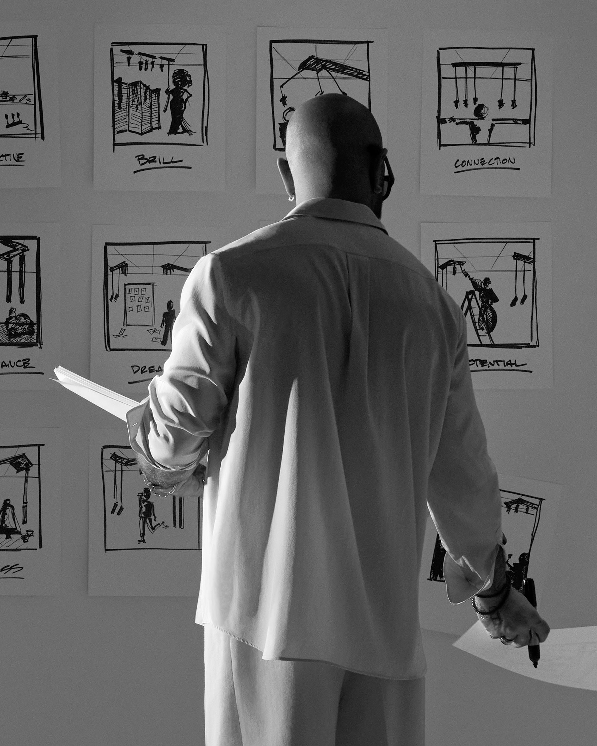 Black and white image of a man studying storyboard sketches pinned on a wall while holding papers and a pen