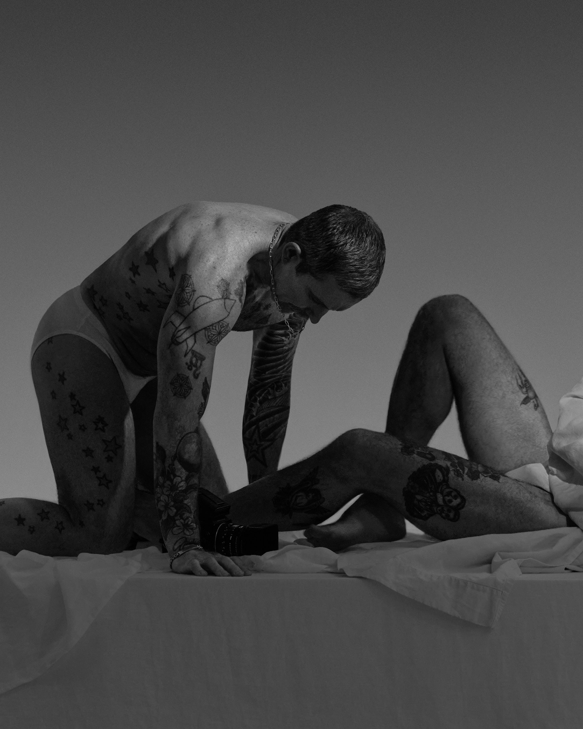Black and white photo of two tattooed men wearing underwear, one kneeling on a bed while the other lies down with legs bent