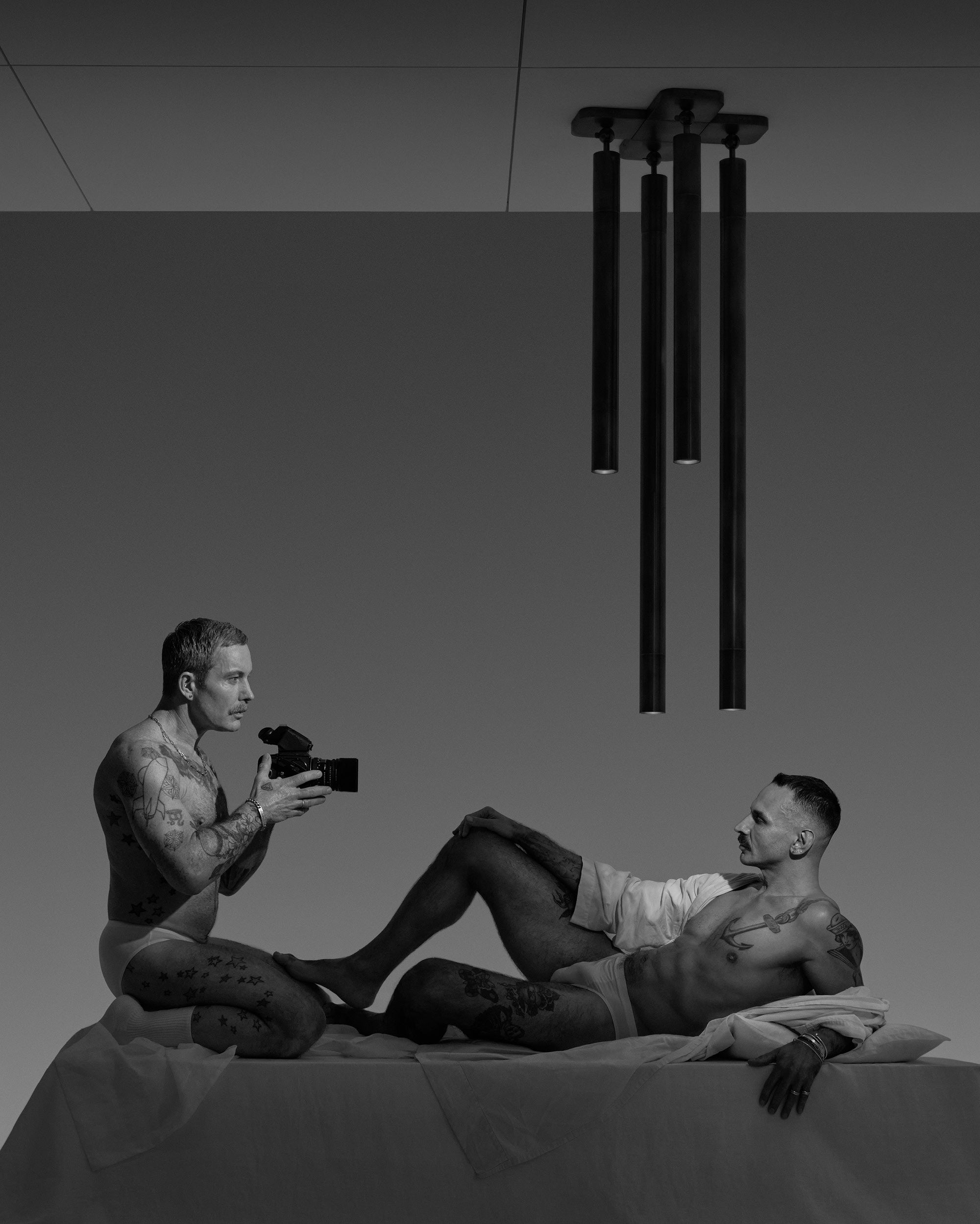 Black and white photo of two tattooed men in underwear, one holding a camera and the other reclining on a bed under pendant lights.