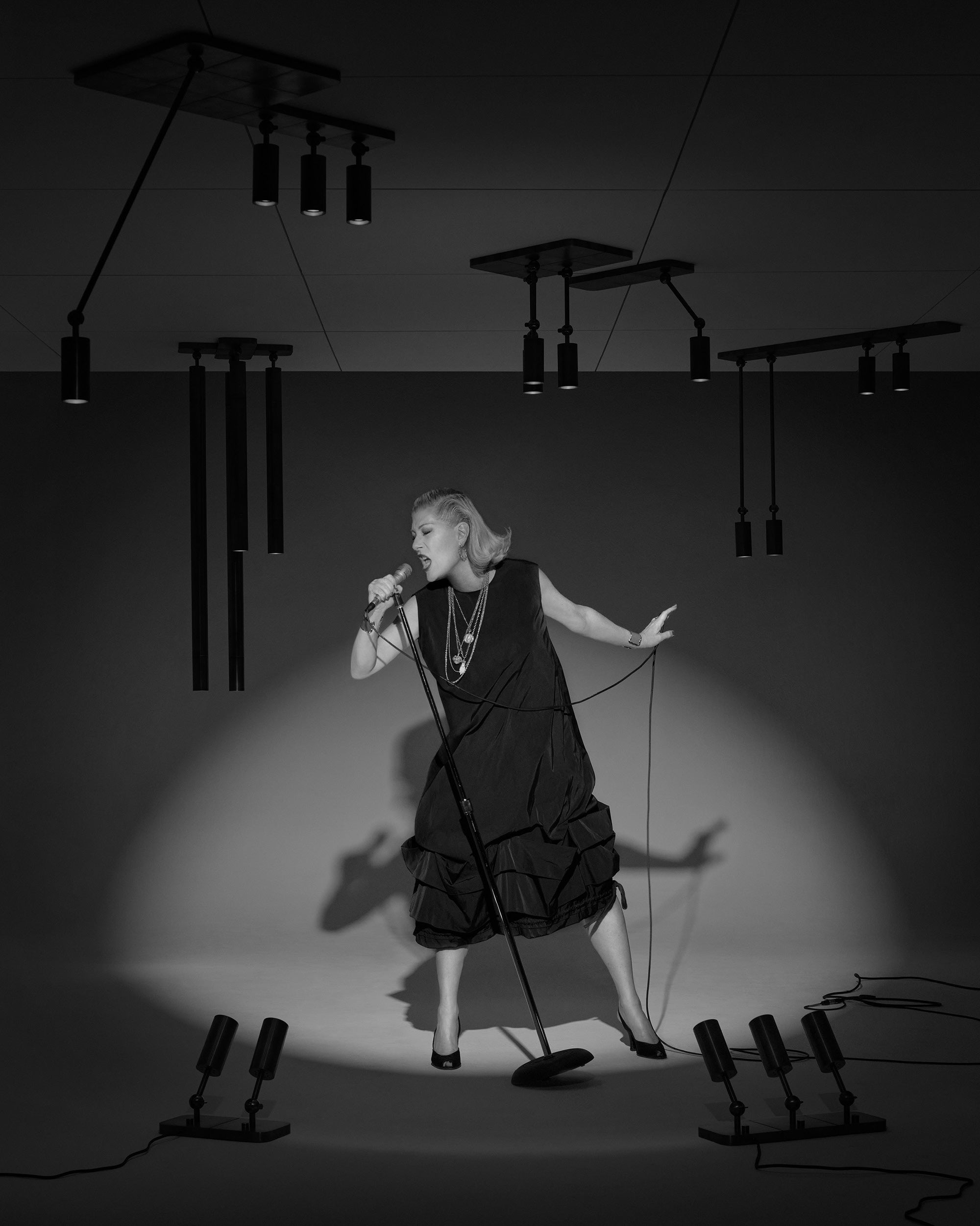 Black and white photo of a woman in a black dress singing into a microphone with dramatic spotlight and overhead lights