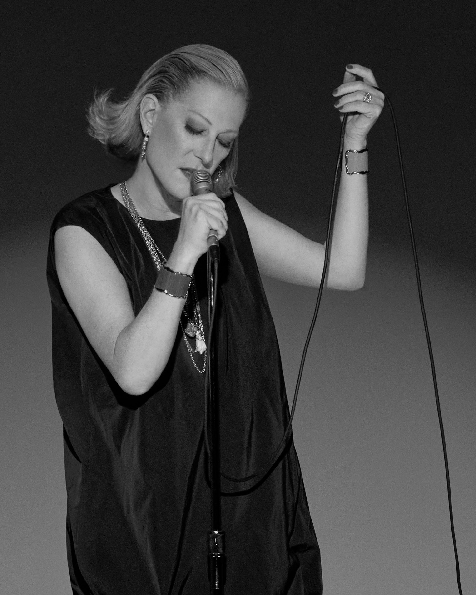 Black and white photo of a woman with slicked-back hair holding a microphone and singing with closed eyes against a plain backdrop