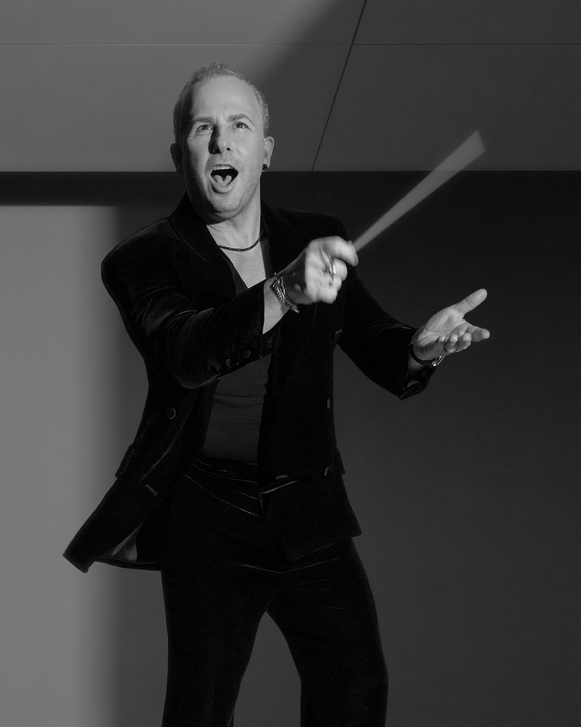 Black and white portrait of a man in a velvet suit conducting with a baton against a minimalist background