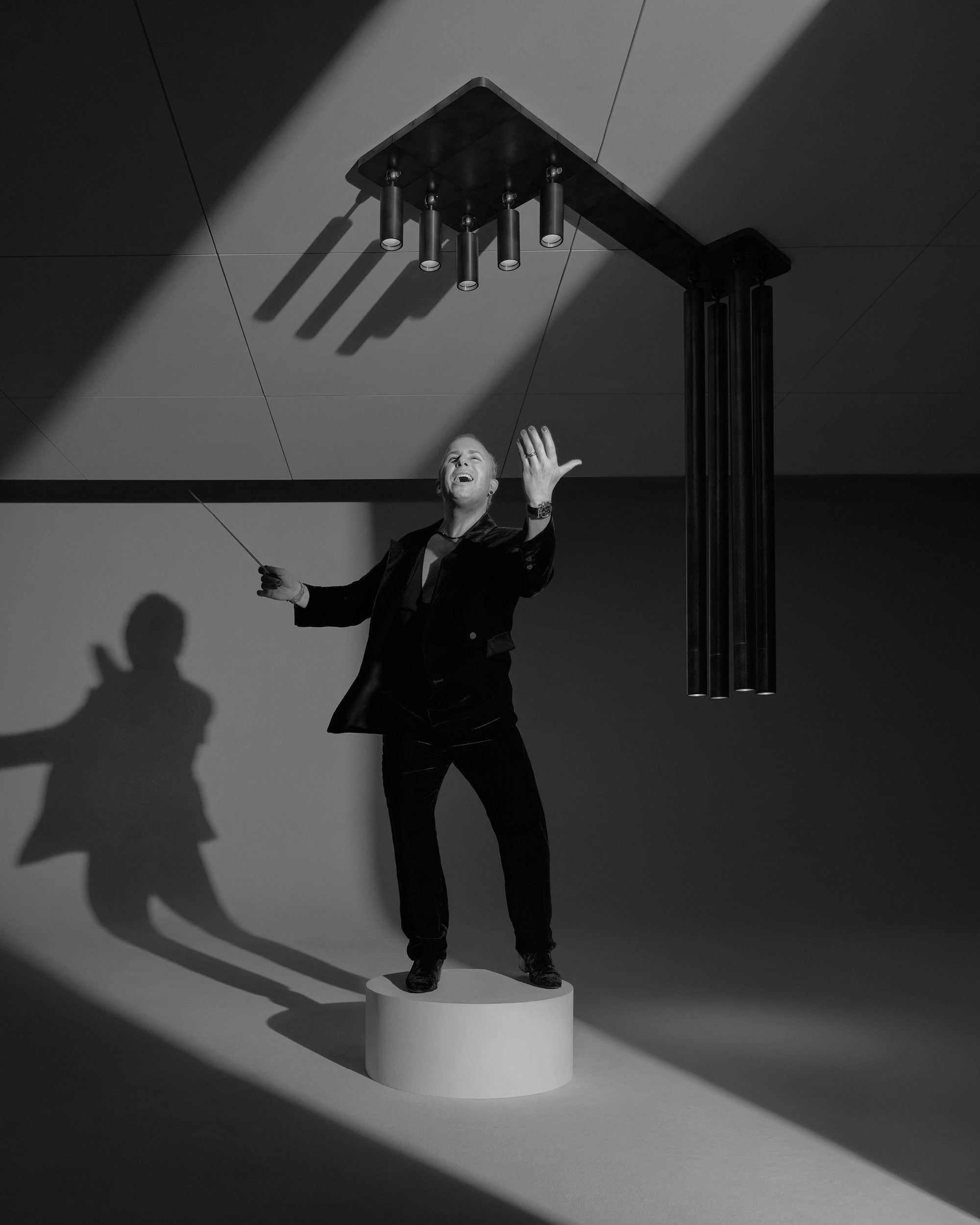 Black and white image of a man in a suit standing on a round pedestal under a modern ceiling light casting shadows