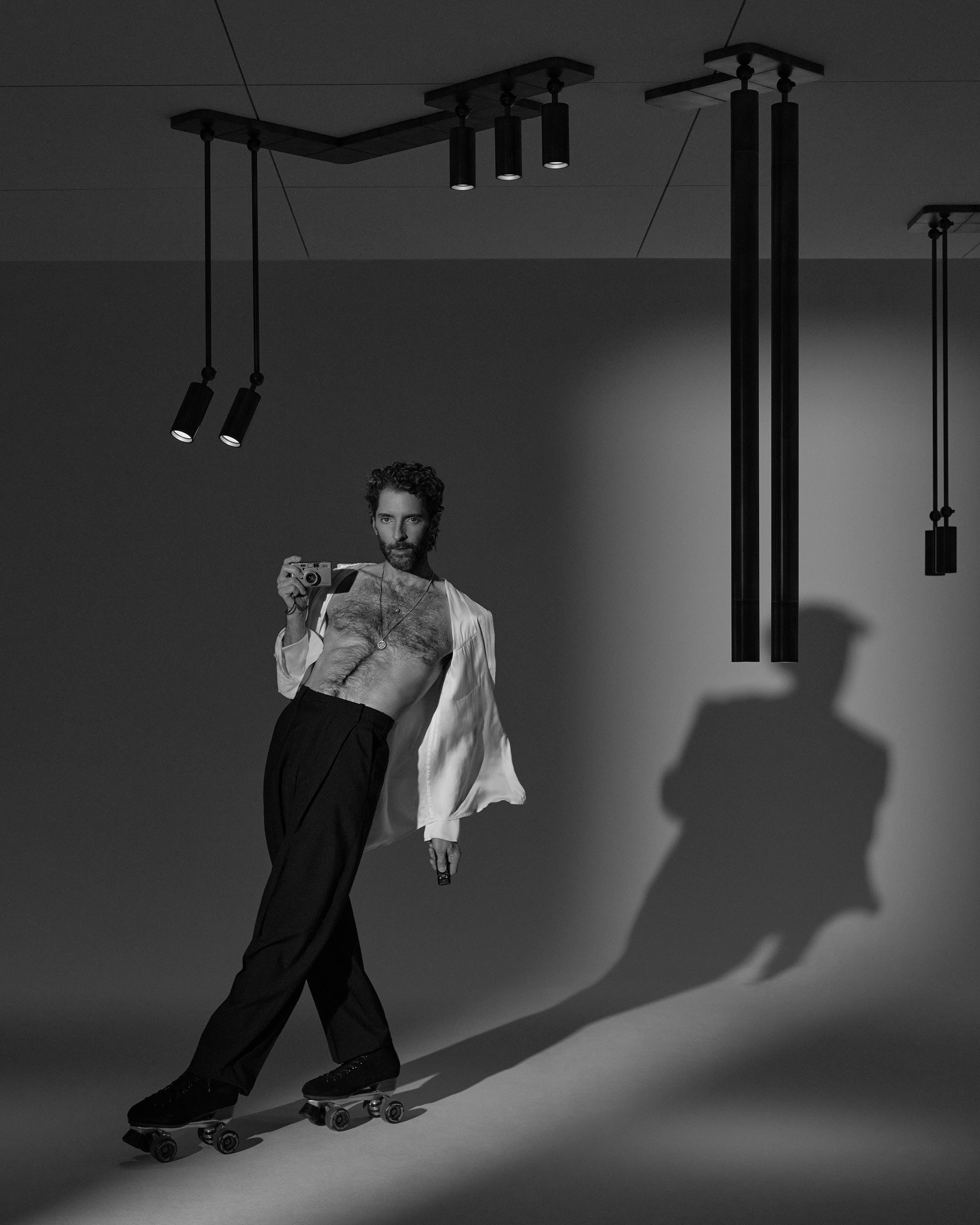 Black and white image of a shirtless man in wide pants and an open shirt roller skating indoors under ceiling spotlights