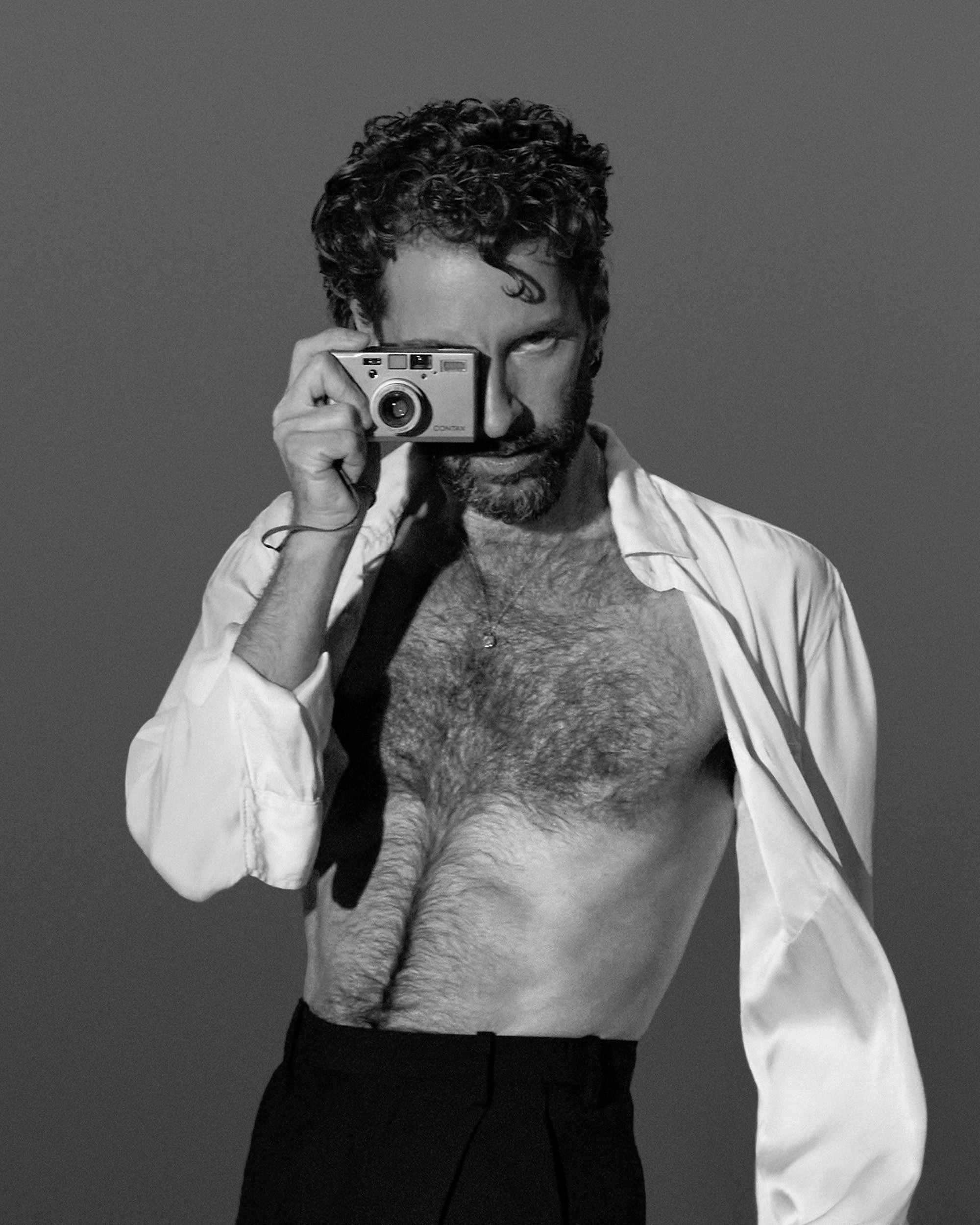 Black and white portrait of a shirtless man with curly hair holding a vintage camera up to his eye against a plain background