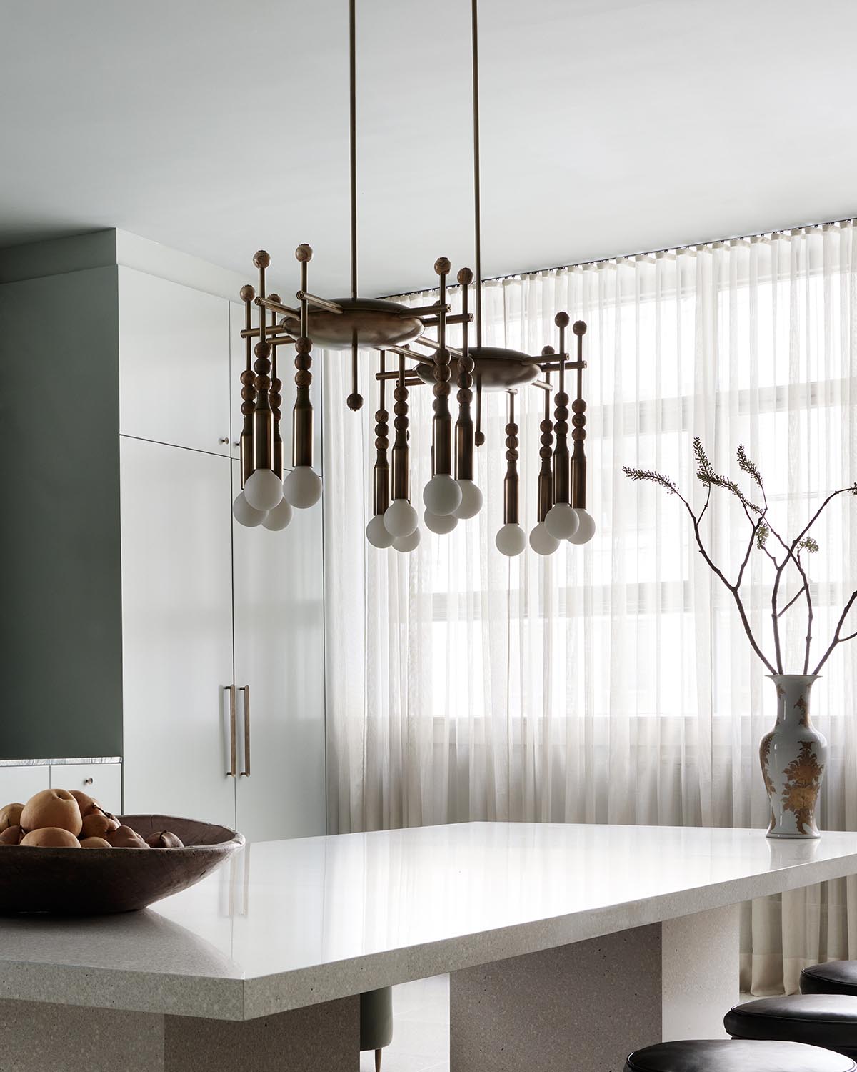 Modern brass pendant light fixture with spherical bulbs above white stone kitchen island and decorative vase near sheer curtains