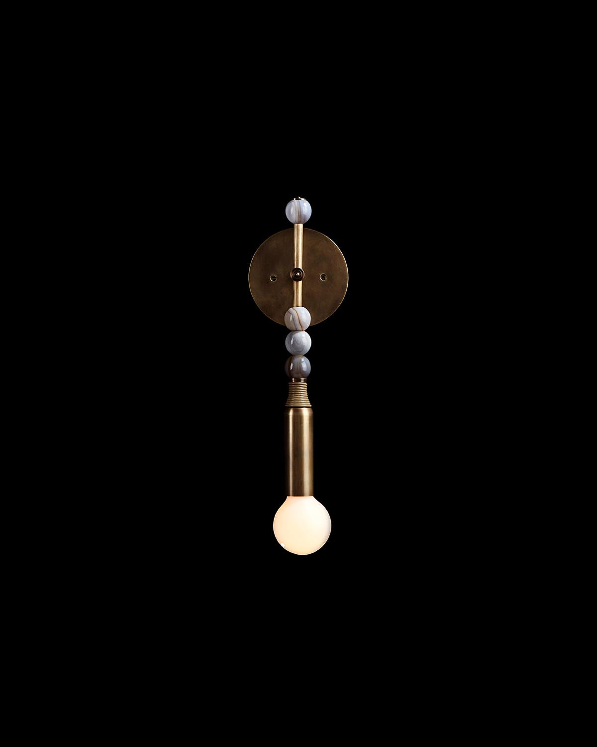 Vertical brass wall sconce with round white bulb and decorative striped stone beads on a black background