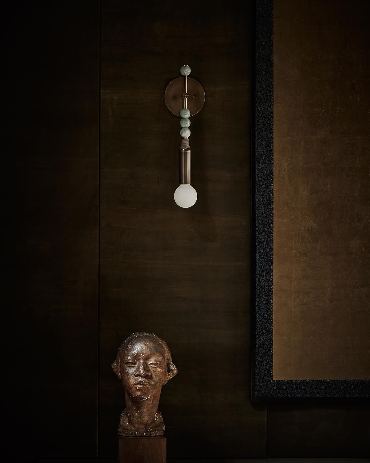 Vertical wall sconce with spherical accents and frosted bulb above bronze sculpted bust on dark textured wall background