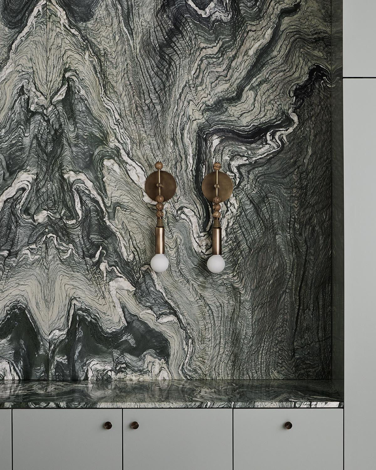 Pair of brass TALISMAN sconces with exposed bulbs mounted on a dark marble veined wall above white cabinetry