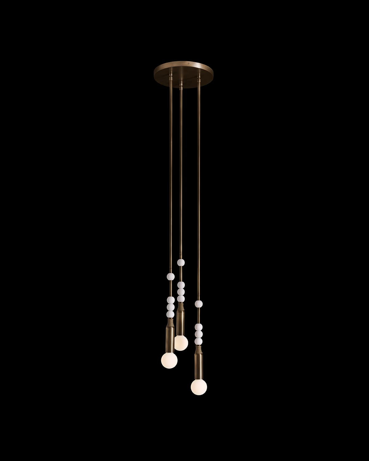 Minimalist pendant light fixture with three suspended bulbs and decorative white beads on brass rods against black background