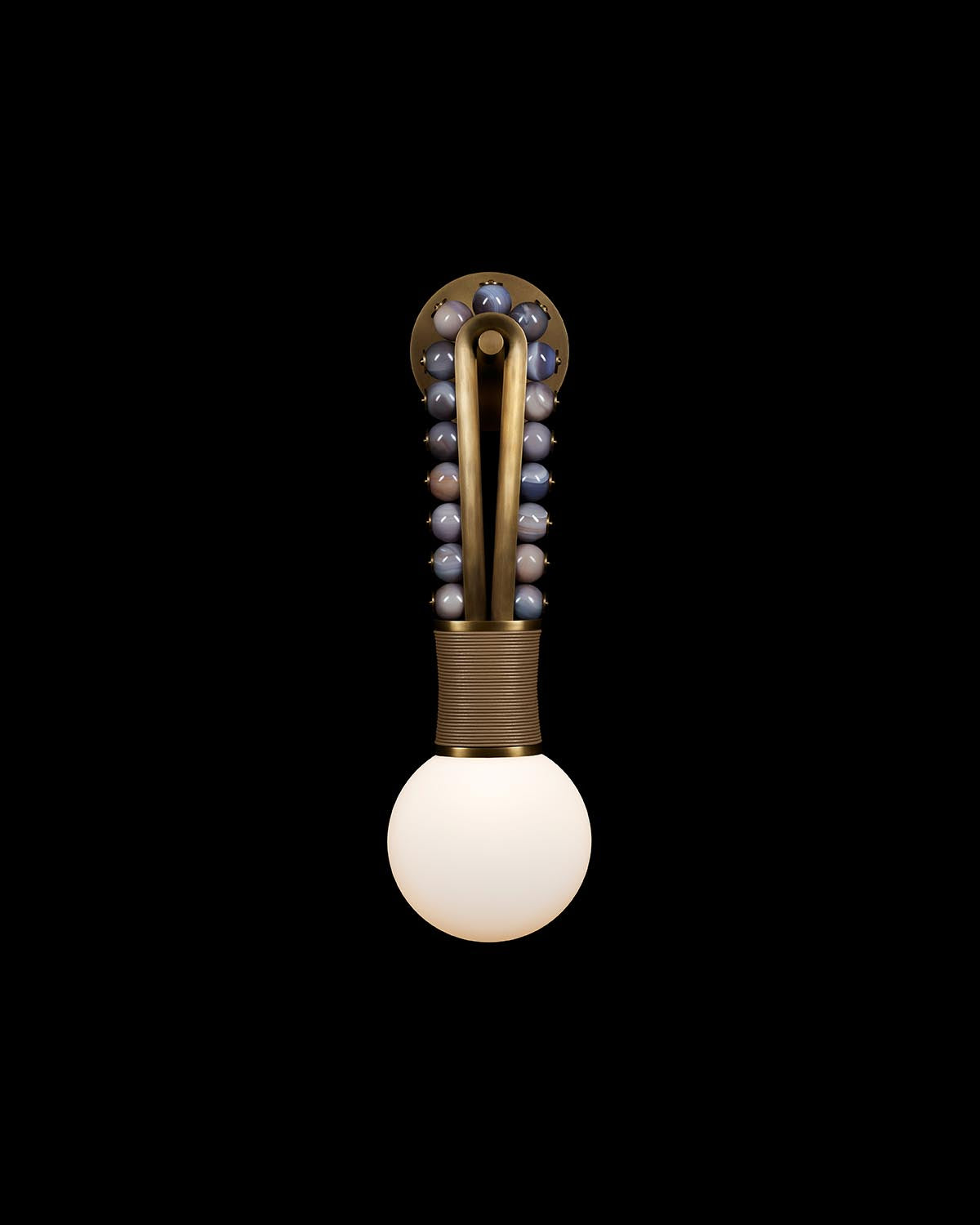 Wall sconce with brass finish, looped vertical arm adorned with gray agate spheres, and illuminated frosted white globe light