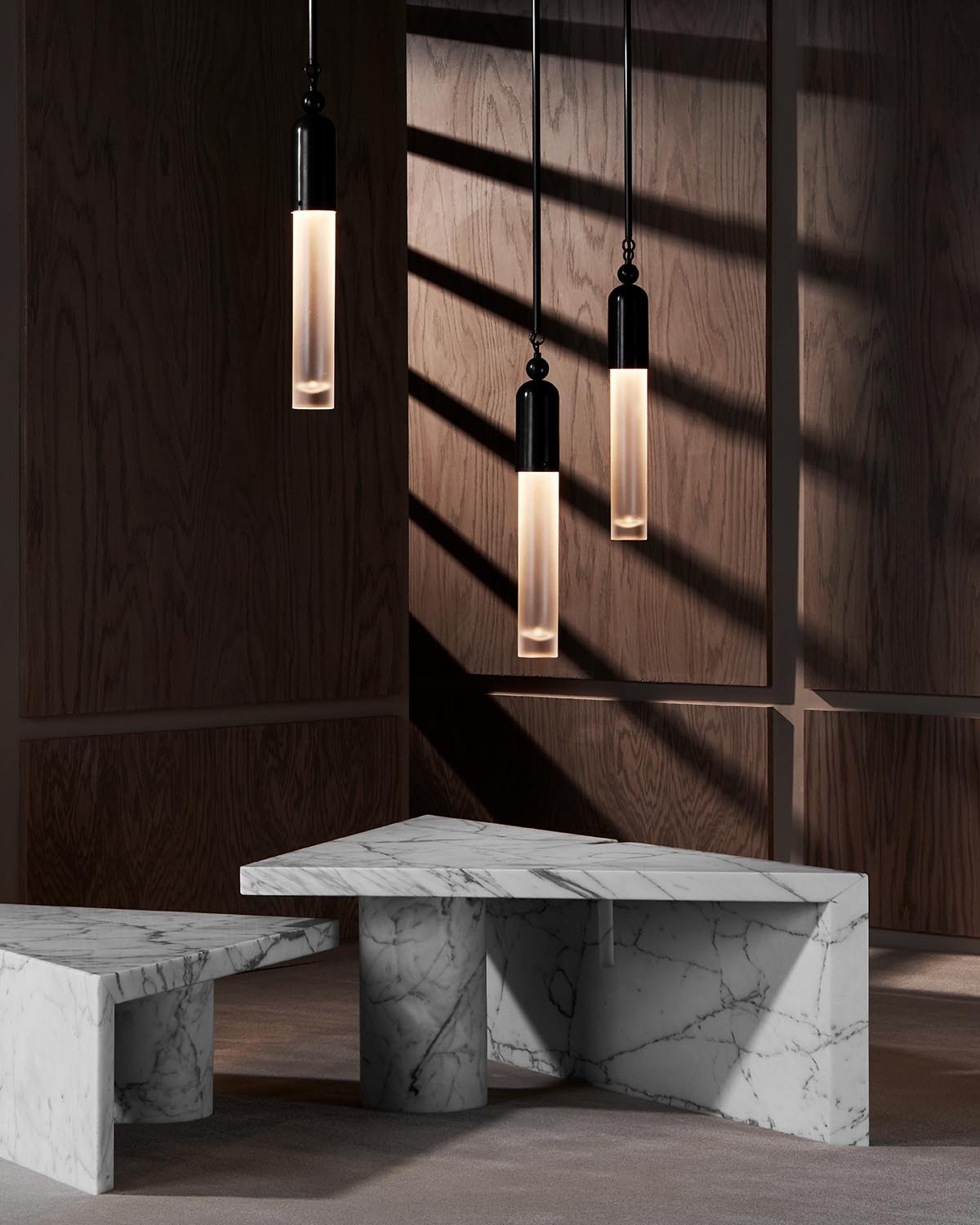 Three cylindrical pendant lights with glowing frosted glass and black tops hanging above white marble tables in a dark wood room