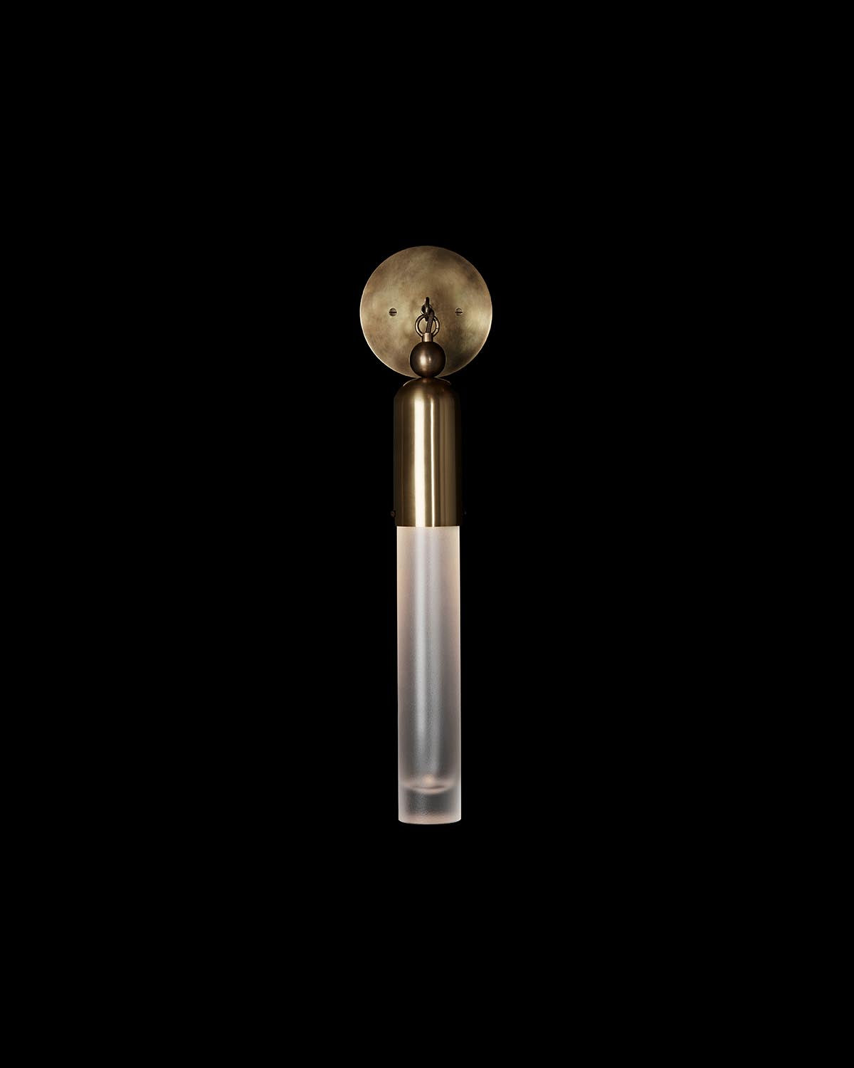 Minimalist wall sconce with round aged brass backplate and frosted cylindrical glass shade against black background