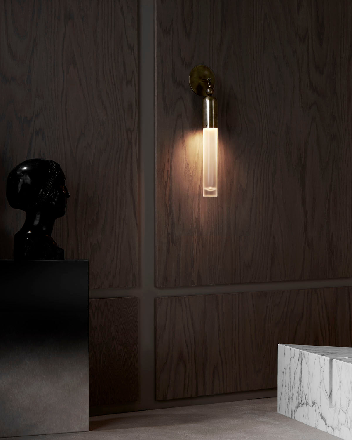 Minimalist wall sconce with frosted glass tube and brass fixture mounted on dark wood paneling in modern interior