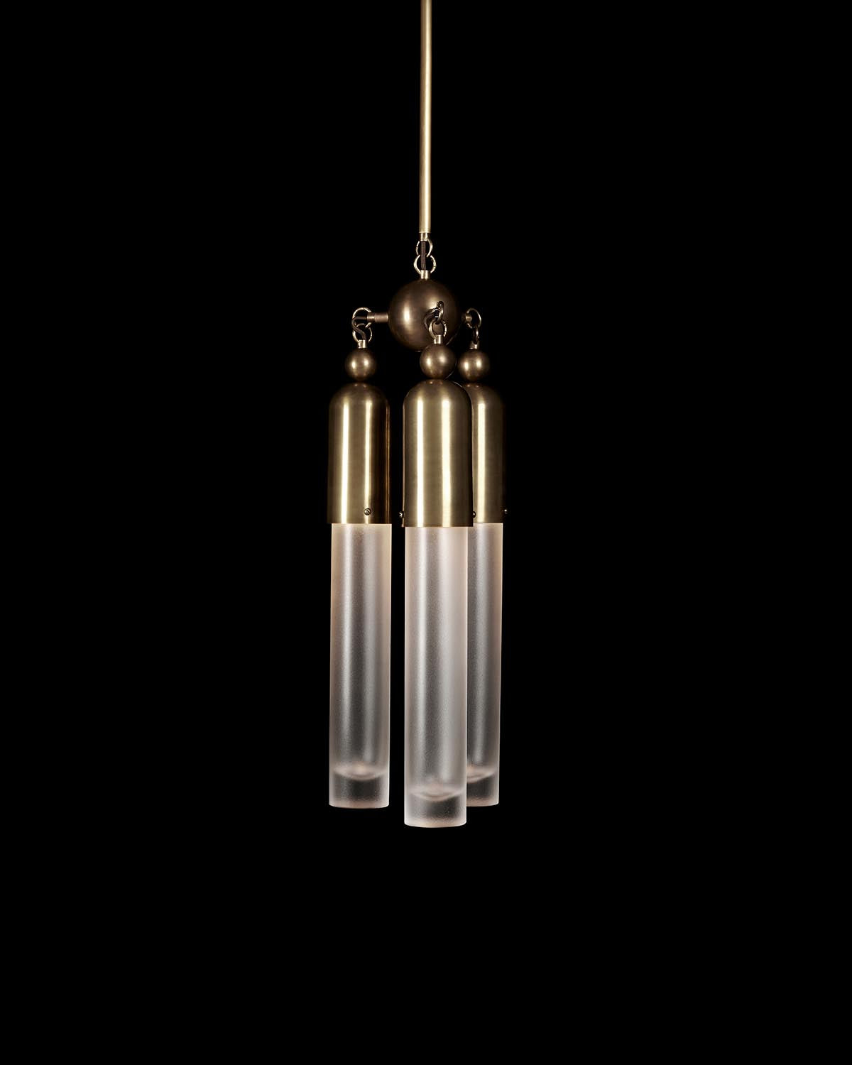 Close-up of a triple pendant light fixture with frosted glass tubes and brushed brass fittings against a black background