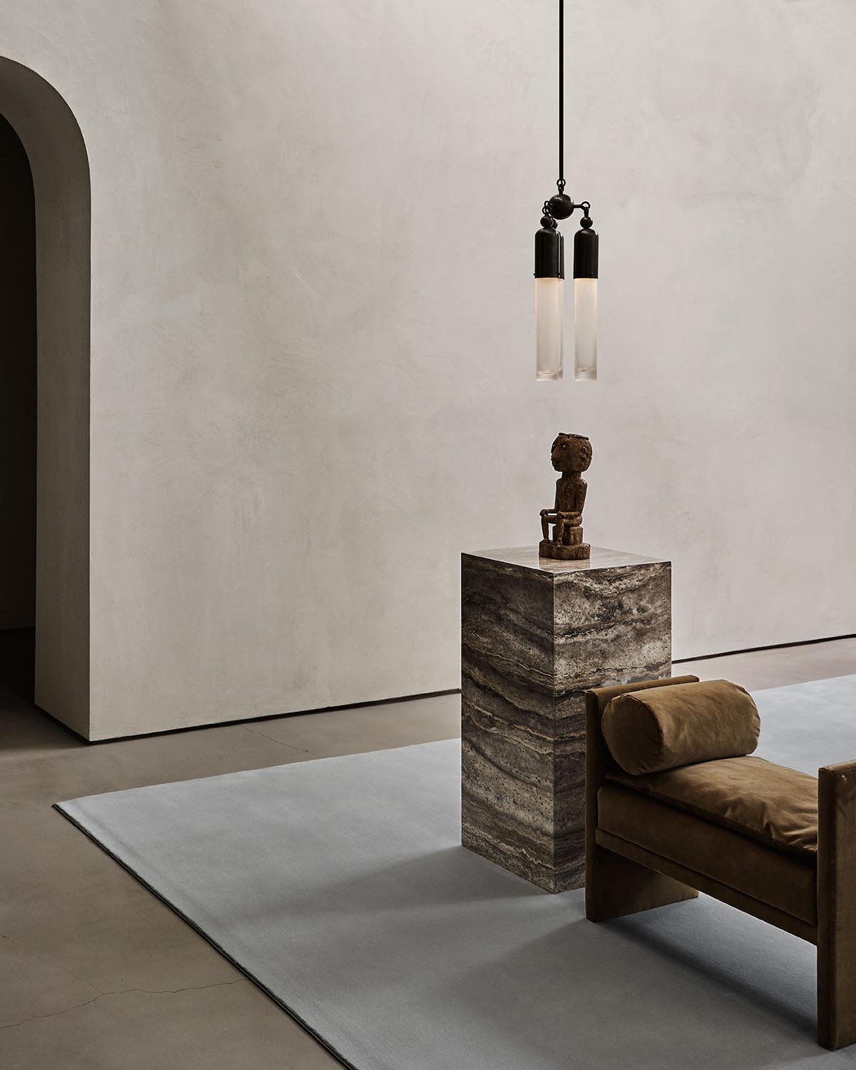 Minimalist interior with three slender pendant lights above a textured stone pedestal holding an abstract wooden sculpture