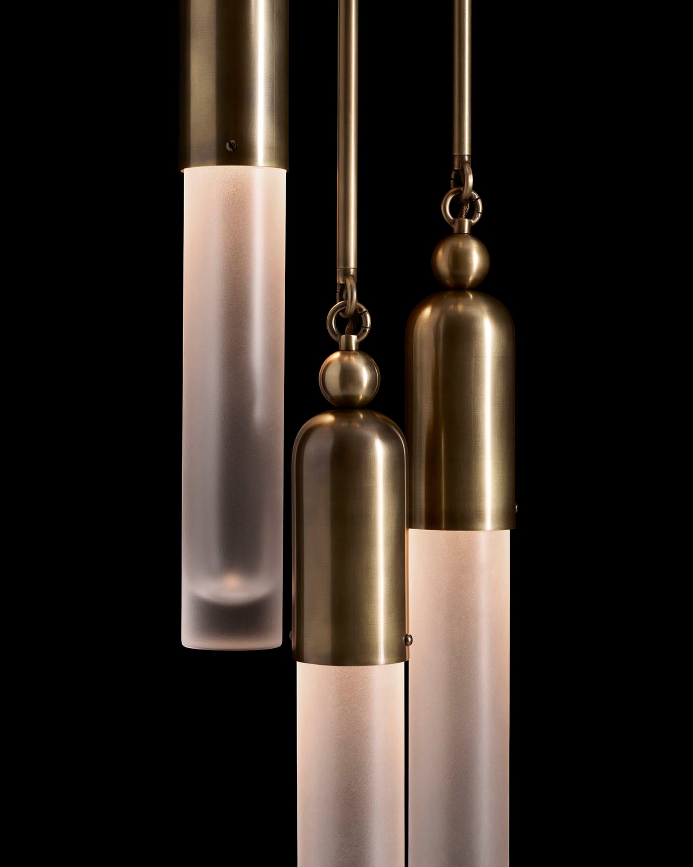 Close-up of three cylindrical pendant lights with frosted glass and brushed brass fittings hanging against a black background