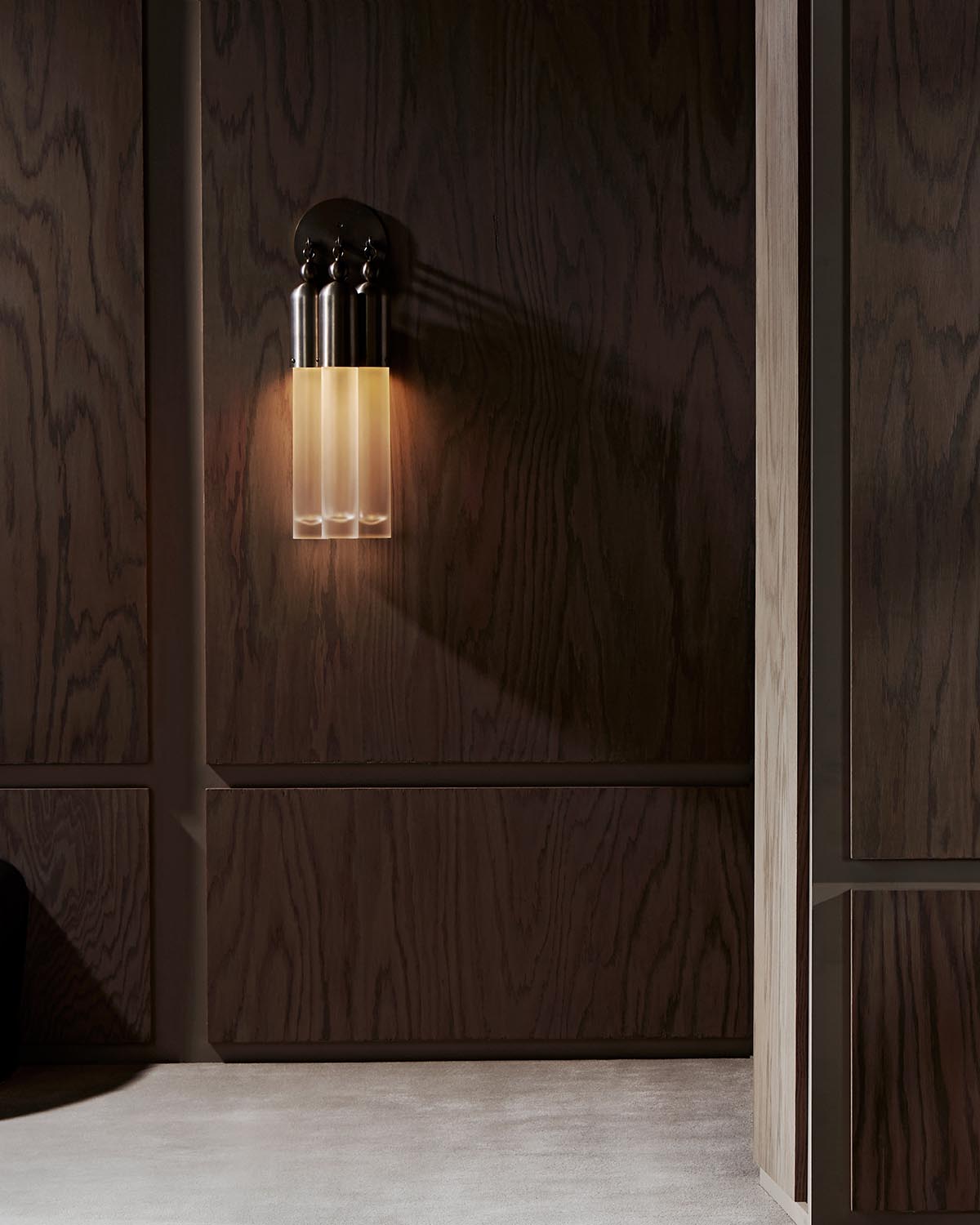 Modern wall sconce with three frosted glass tubes and dark metal base mounted on textured wooden panel wall