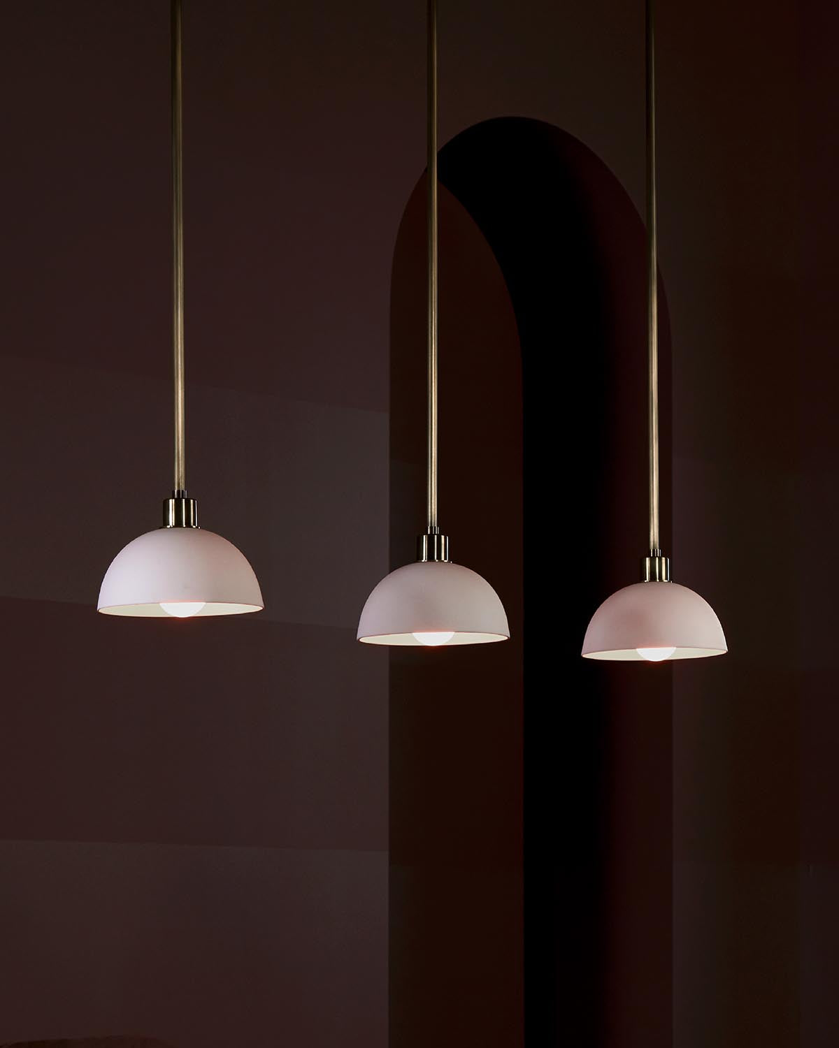 Three white dome pendant lights hanging against a dark brown wall with an arched shadow in the background