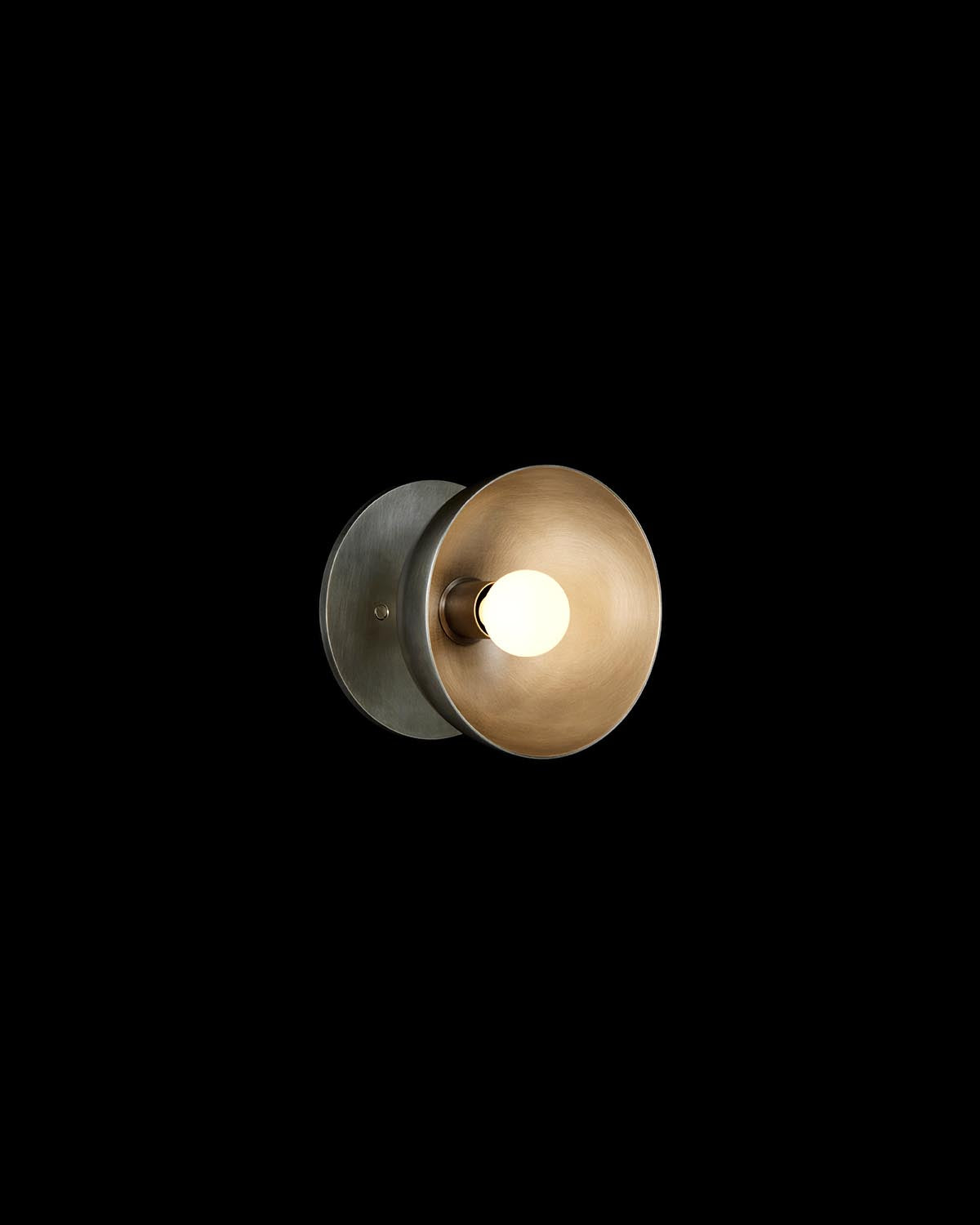 Close-up of a metal wall light fixture with a circular bronze reflector and exposed bulb on a black background