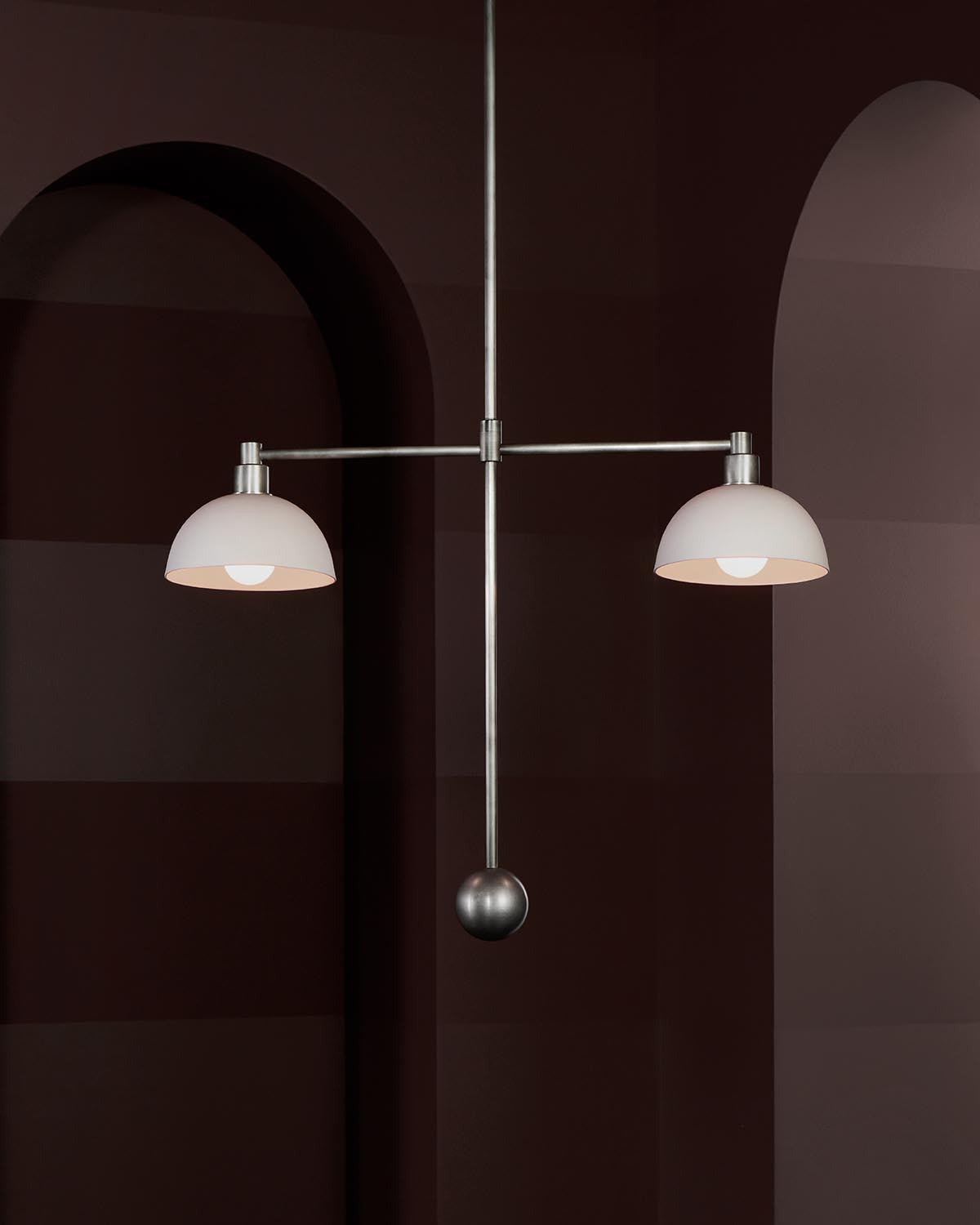 Modern pendant light with two white dome shades and brushed metal frame against a dark arched wall background