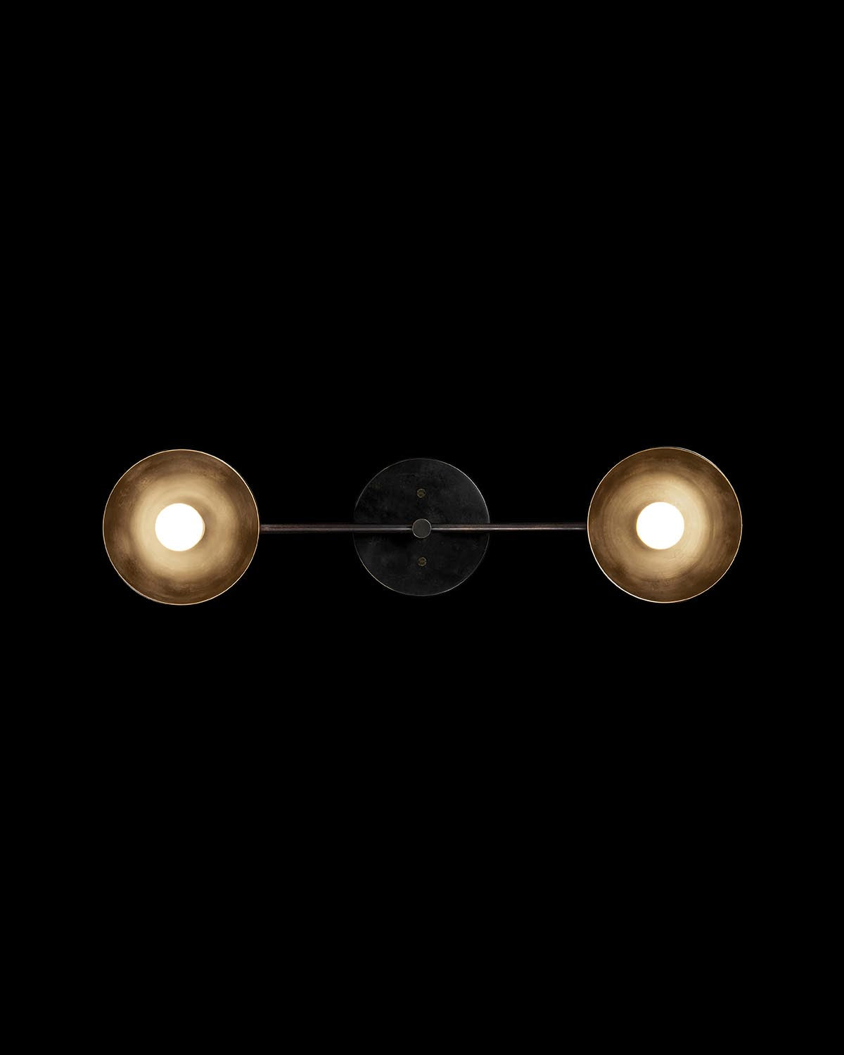Minimalist wall-mounted light fixture with two round brass lampshades connected by a slim dark metal bar on black background