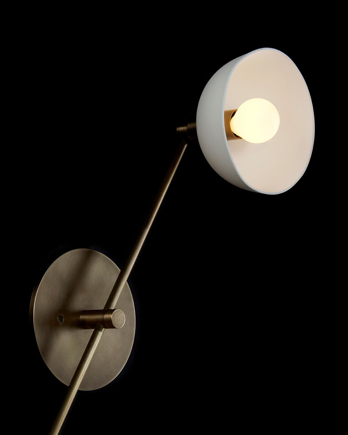Minimalist wall lamp with white conical shade and brass arm mounted on a circular brass base against black background
