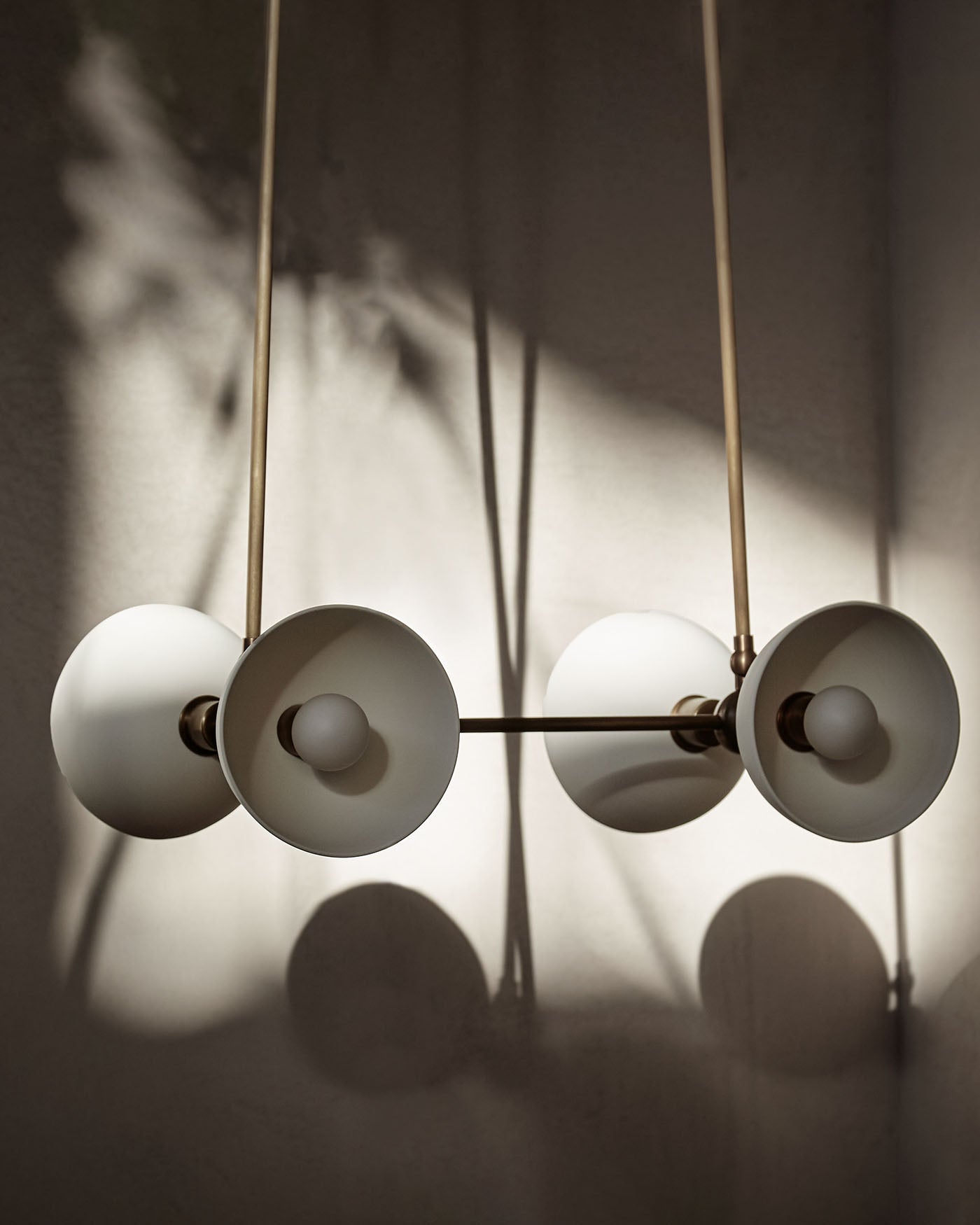 Minimalist trapeze-style pendant light fixture with four matte white shades and brass rods casting soft shadows on wall
