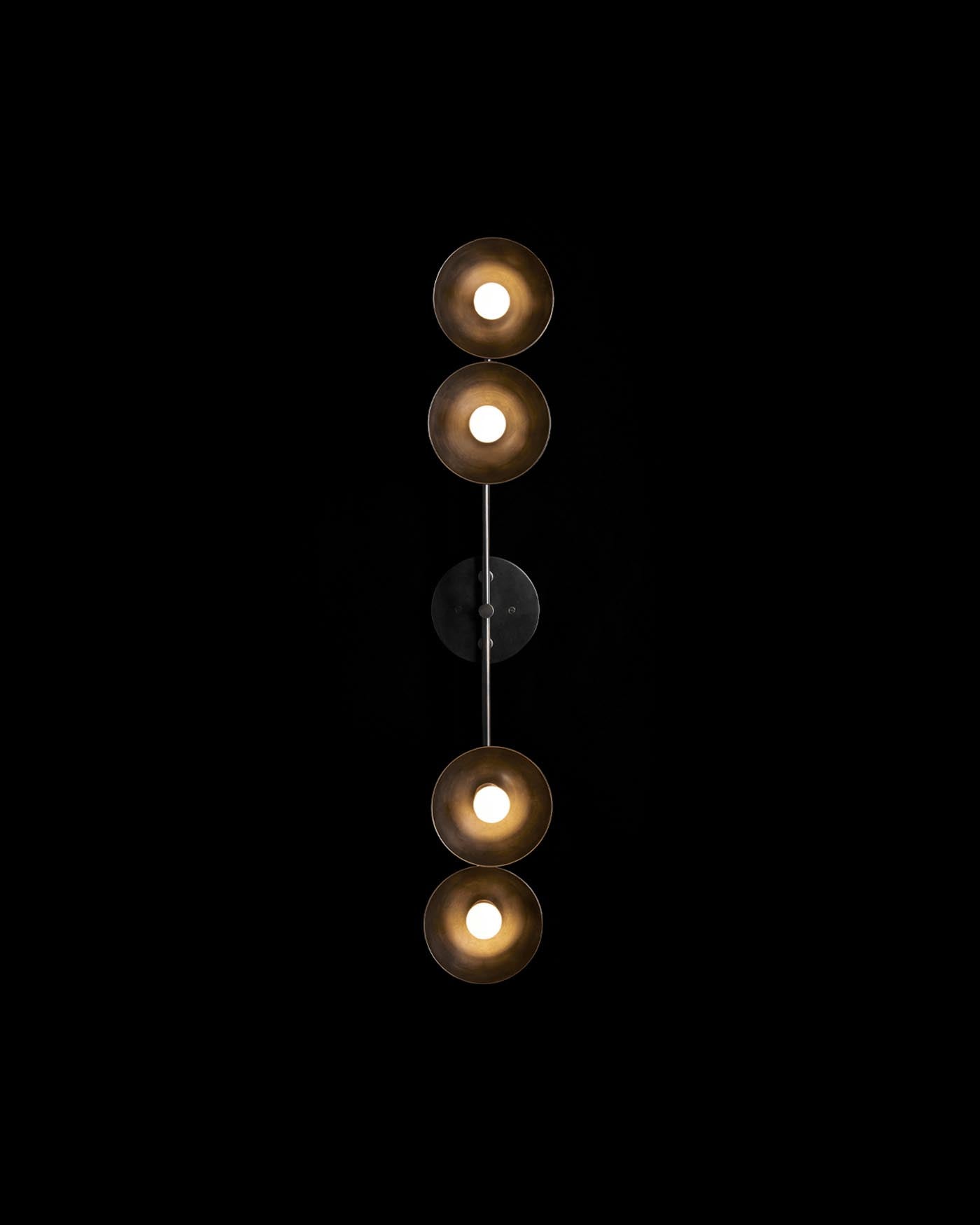 Vertical wall light fixture with four circular lampshades in a brass finish against a black background
