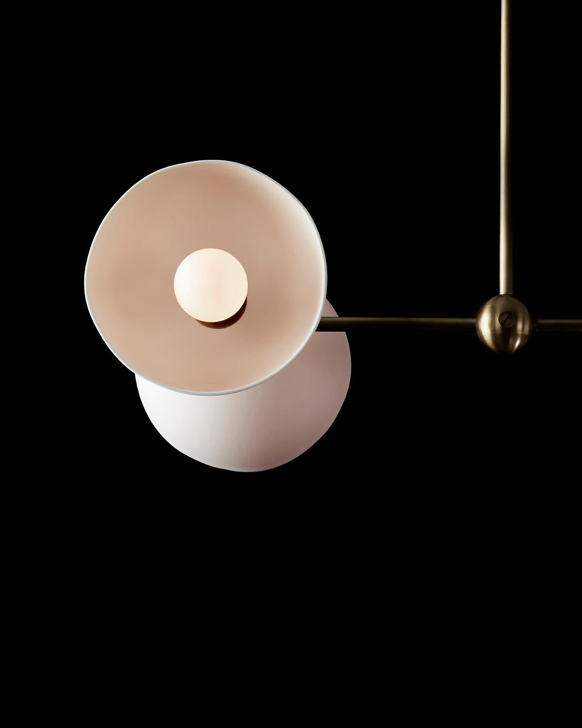 Close-up of a modern pendant light with two overlapping curved shades and central glowing bulb on a brass rod against black background