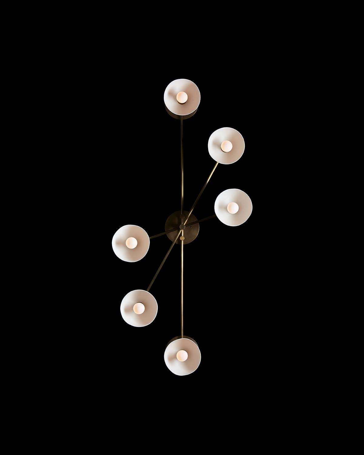 Overhead view of a modern chandelier with six round white lampshades on slender brass arms against a black background
