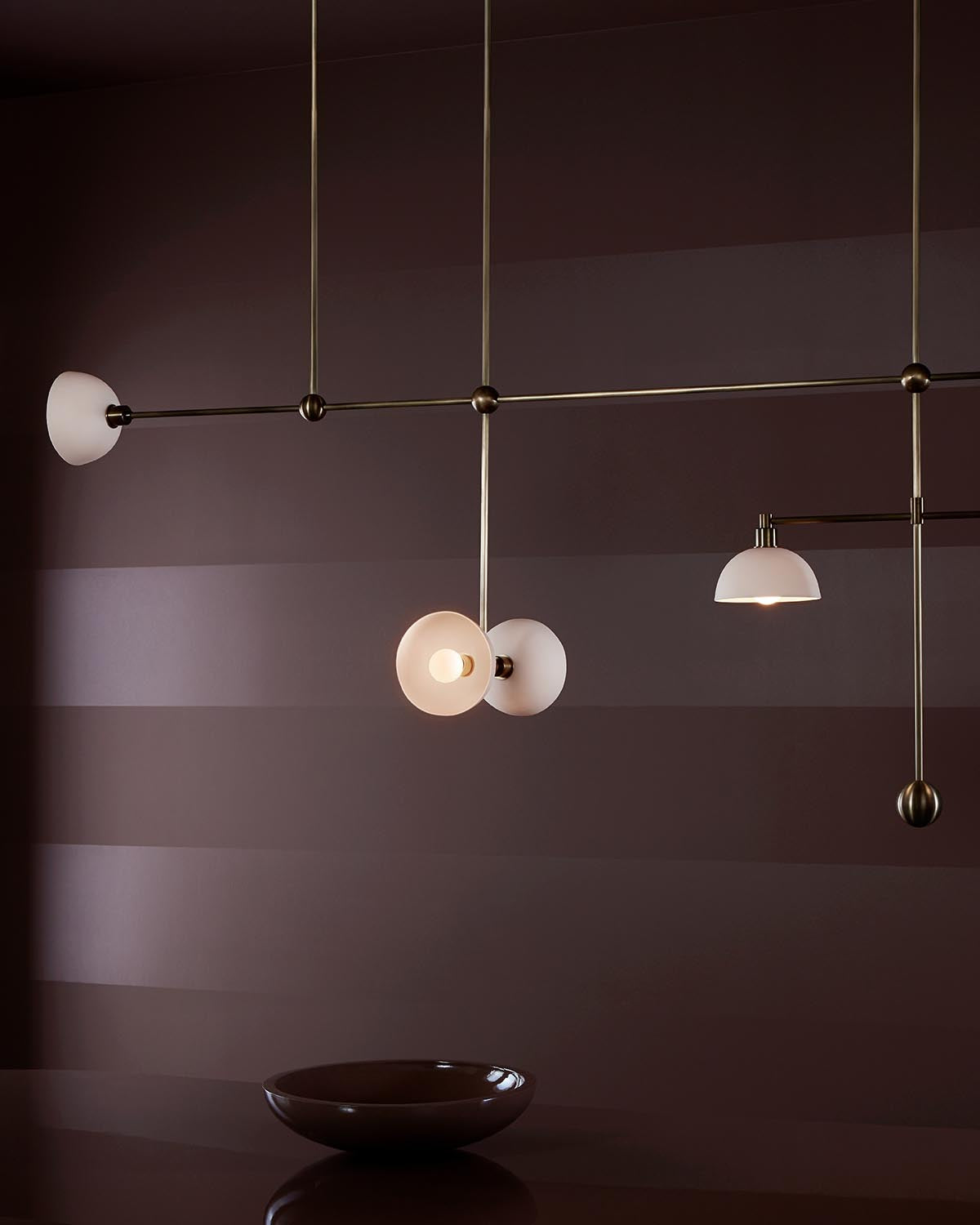 Minimalist trapeze-style ceiling mobile with multiple round white glass lampshades and brass rods against a dark striped wall