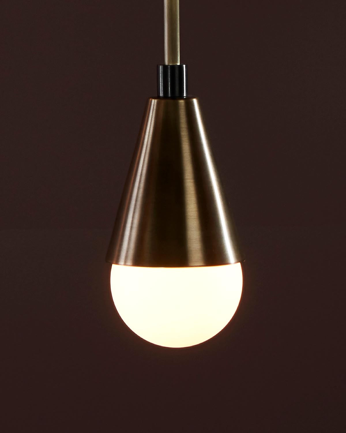 Close-up of a single pendant light with brushed brass conical shade and glowing round bulb against a dark background
