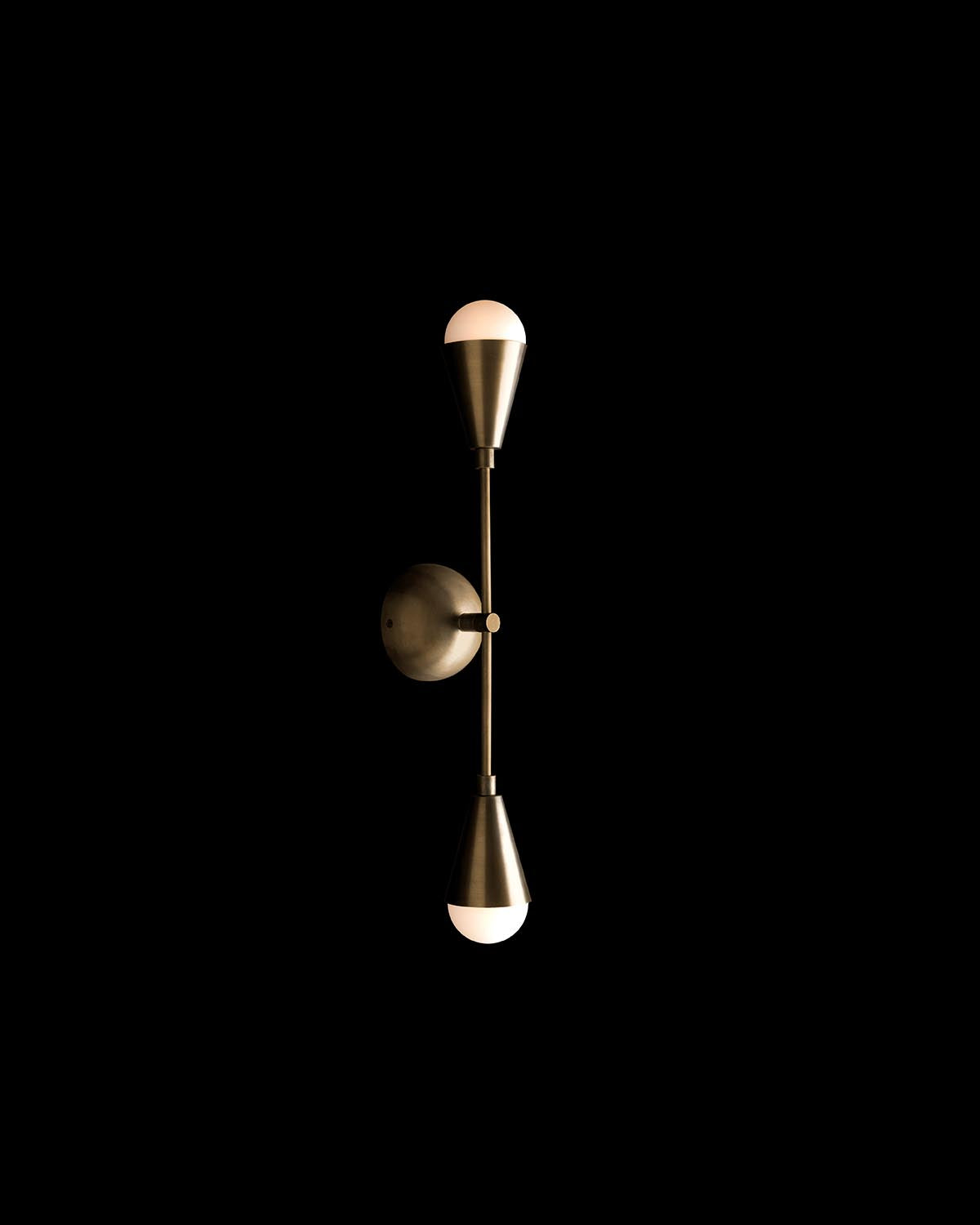 Vertical modern wall sconce with two cone-shaped metallic shades and glowing white bulbs on black background