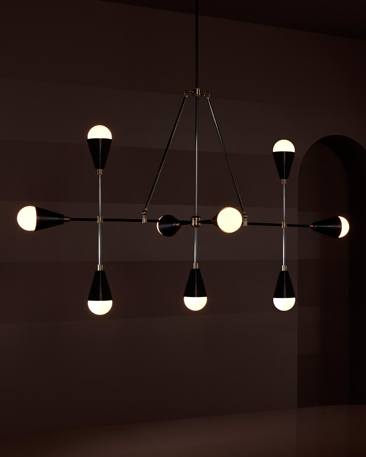 Modern linear pendant light fixture with nine glowing bulbs in black and brass finishes against dark wall