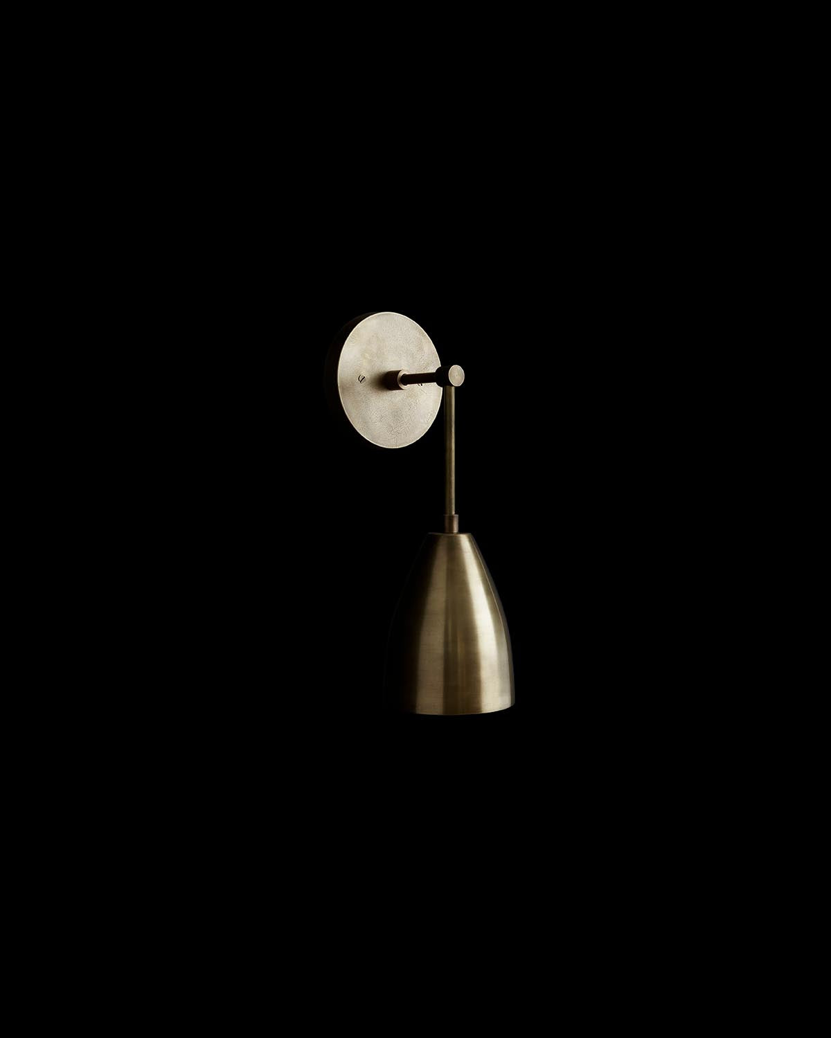 Minimalist brass wall sconce with conical shade and circular mounting plate on a black background