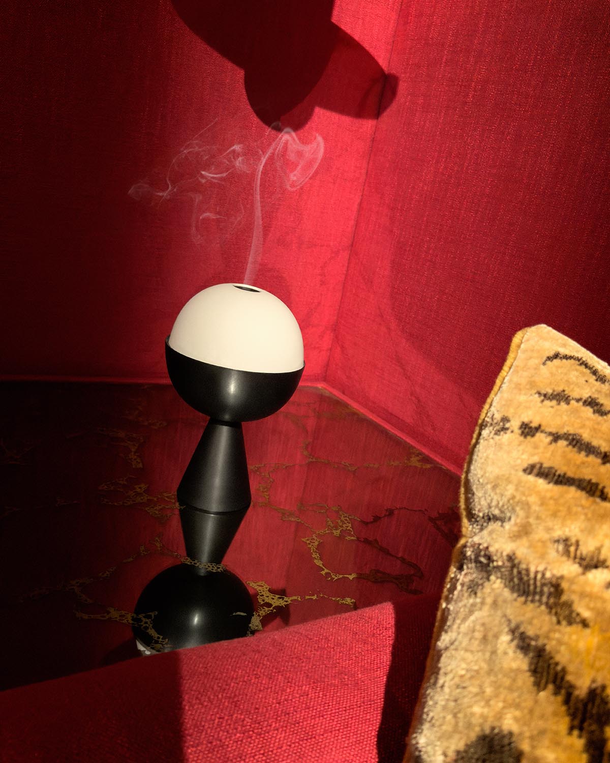 Modern black and white spherical censer emitting smoke on a reflective dark surface with red and patterned fabric background