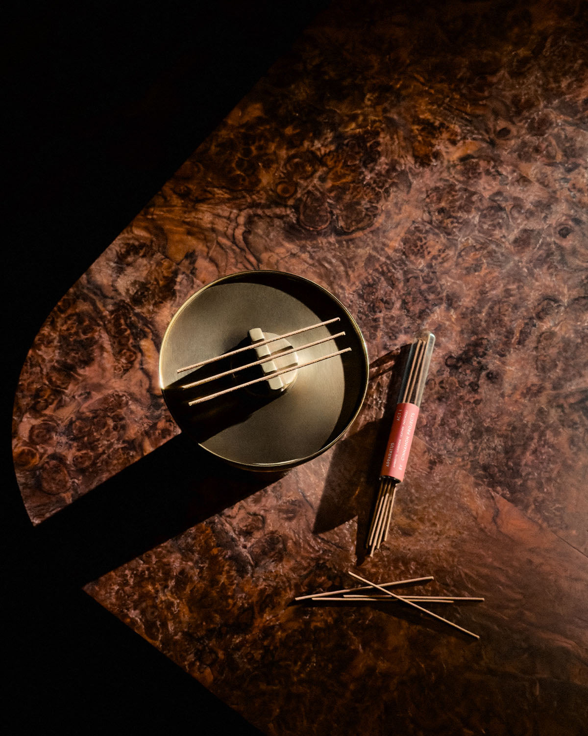 Top view of a dark round censer with incense sticks resting on a textured brown surface with sunlight and shadows
