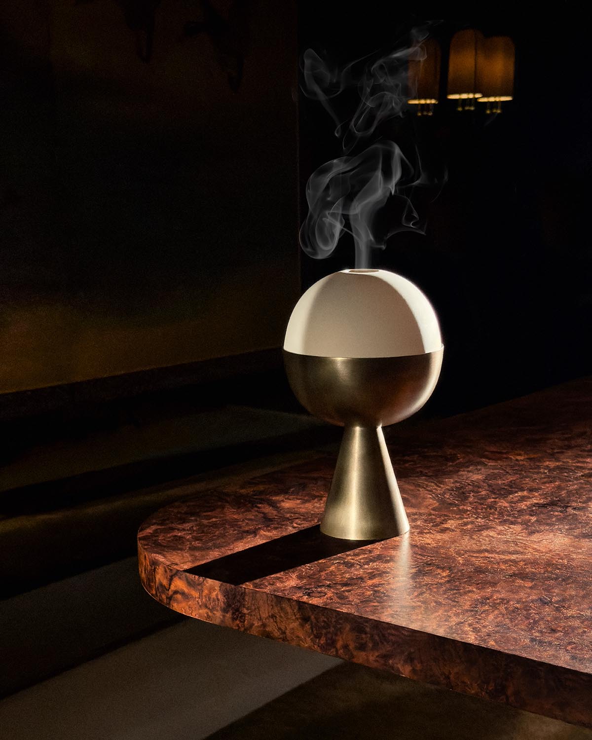 Modern spherical censer with brass base releasing smoke, placed on a textured dark wooden table under warm lighting
