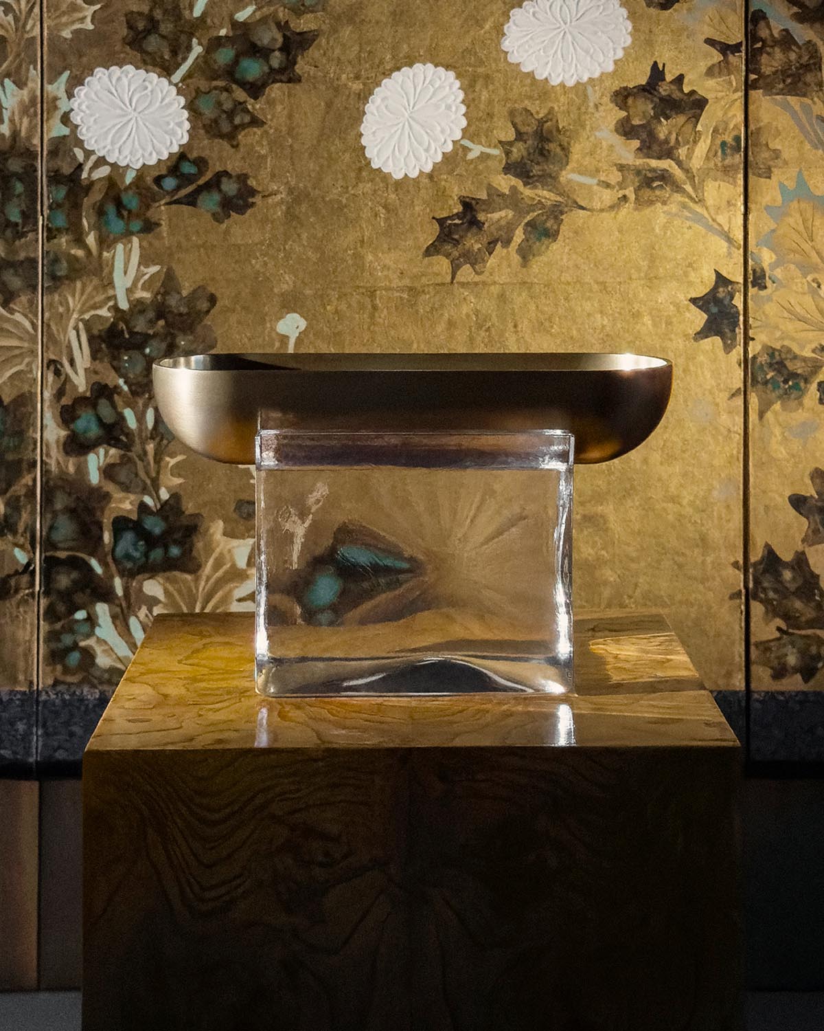 Minimalist bronze vessel resting on a translucent rectangular glass block against a floral gold-leaf backdrop