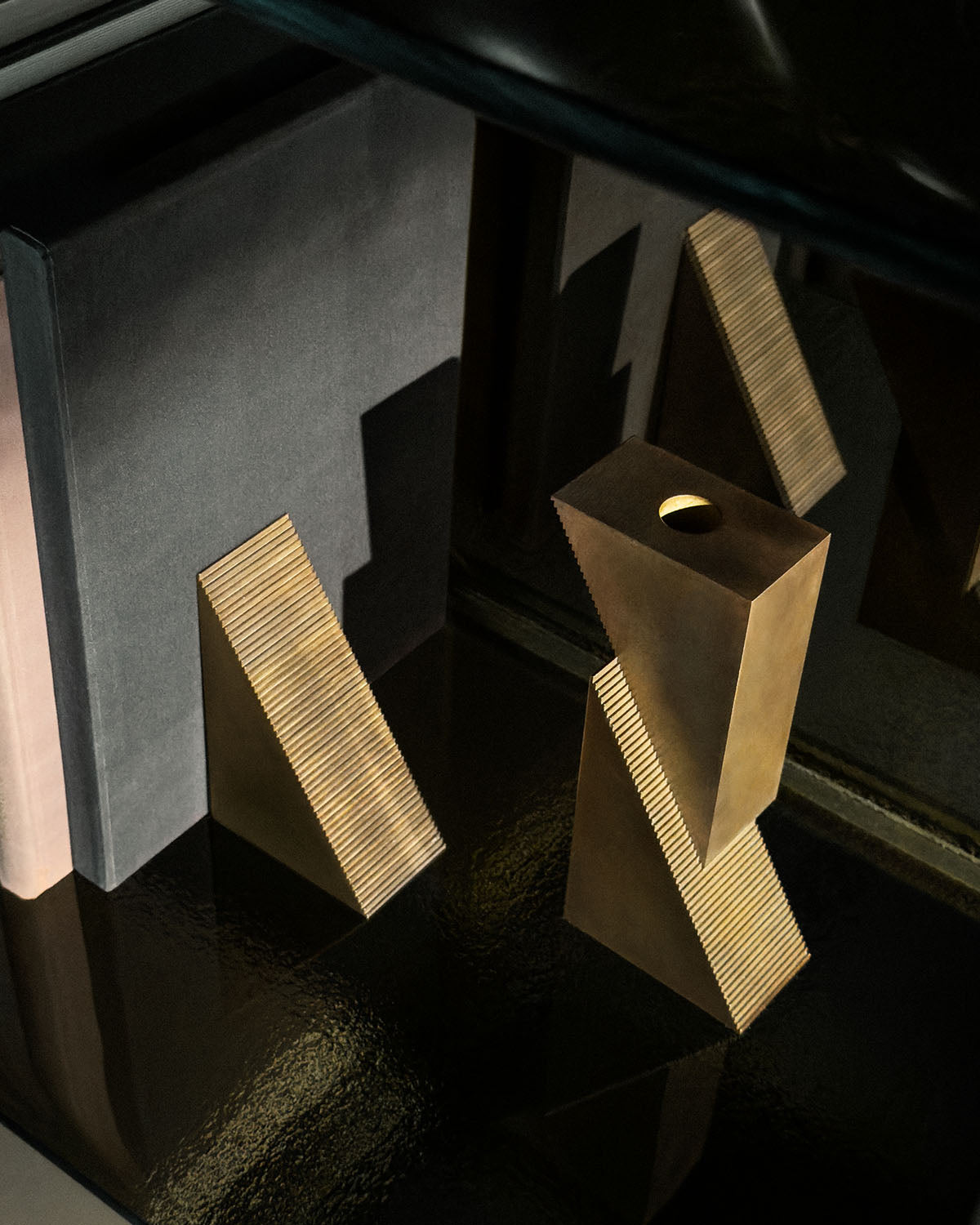 Two geometric brass candle blocks with textured sides reflected on a dark glossy surface against a grey backdrop