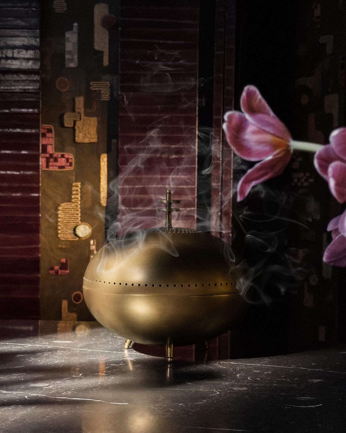 Brass smoking vessel with perforated design emitting smoke, placed on dark marble surface with patterned background and pink flowers nearby