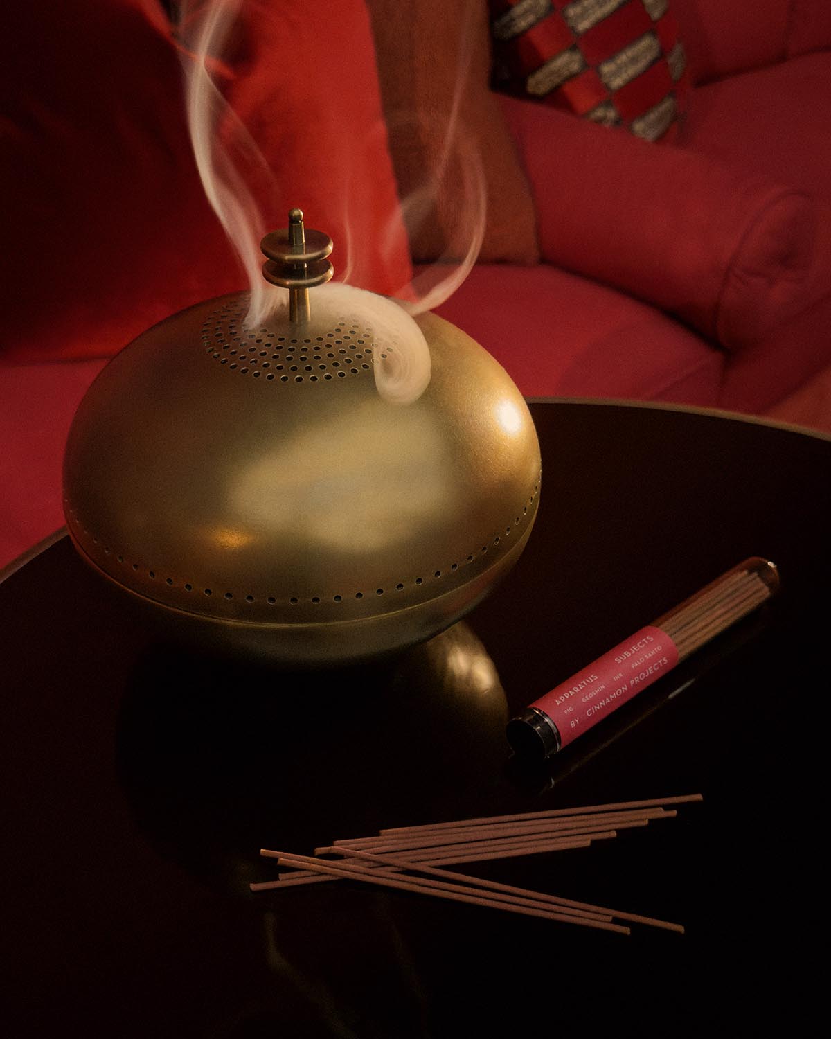 Brass dome-shaped smoking vessel releasing smoke on black table next to incense sticks and red packaging pouch